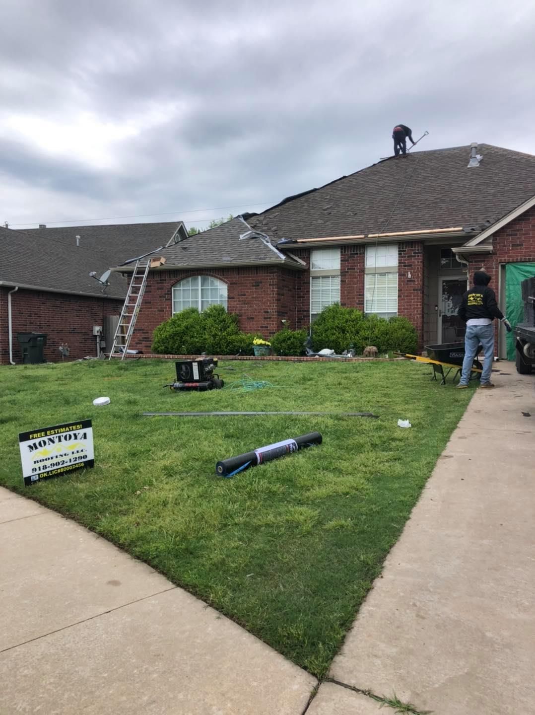 Damaged front lawn and roof of a brick house, with debris and a worker on the roof after a storm.