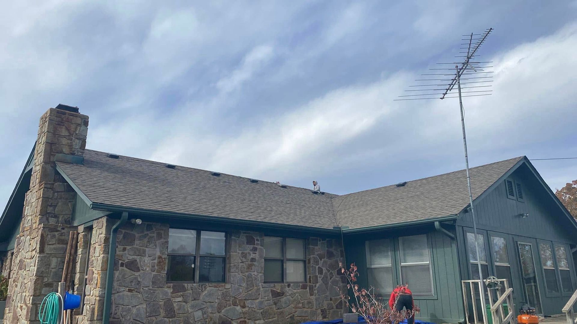 Stone house with a wavy roof, large windows, and a tall antenna under a cloudy sky