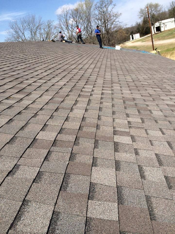 Sloped shingle roof with two people standing at the top near trees and a house
