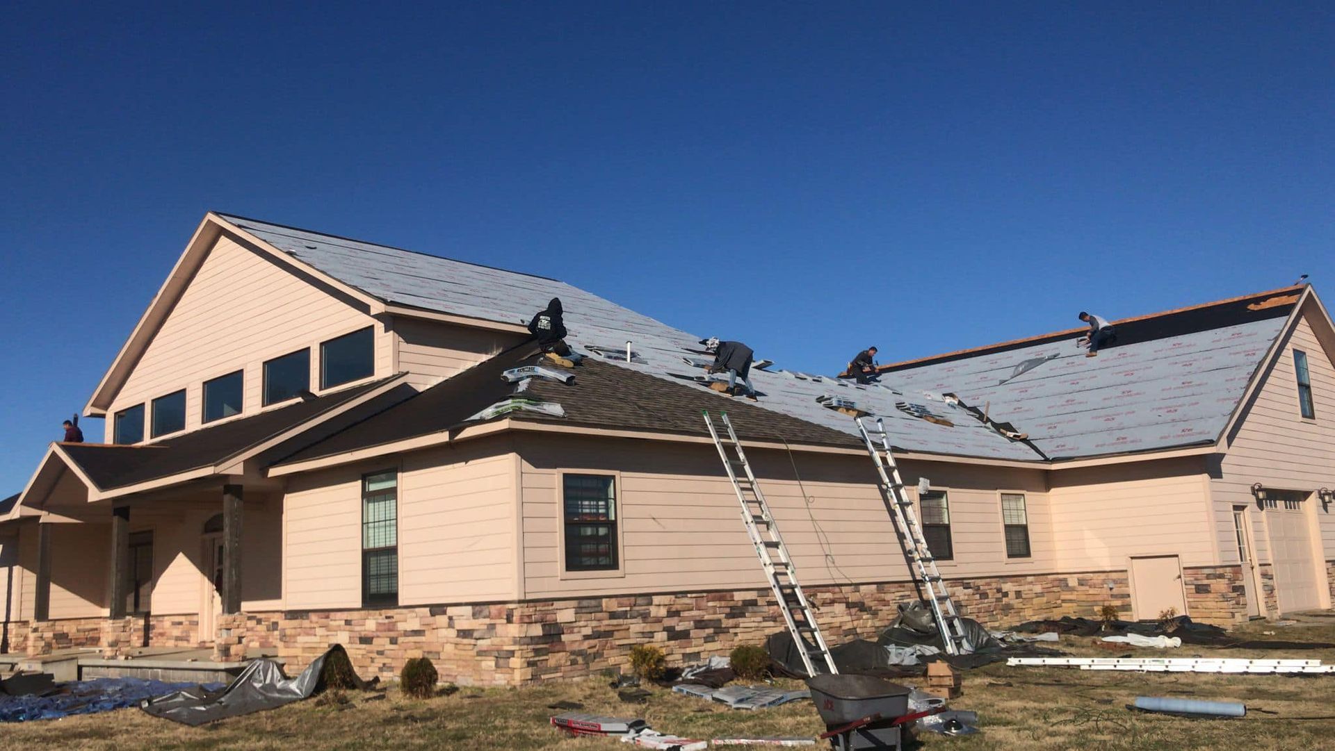 New house under construction with tan siding, gray roof, and ladders on a clear day