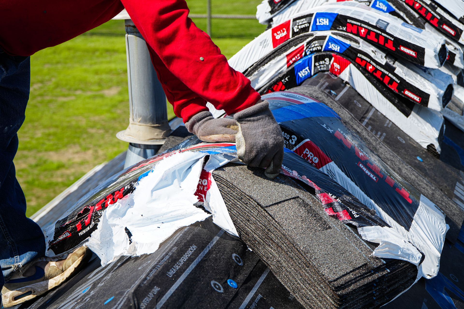 Gloved hands tearing black roofing shingles on a roof, with red sleeves visible