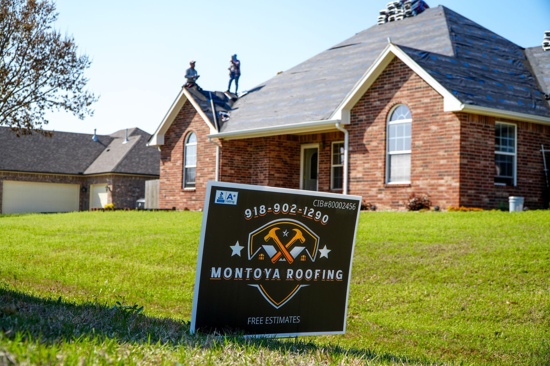 Roofing company yard sign on a lawn in front of a brick house, with workers on the roof