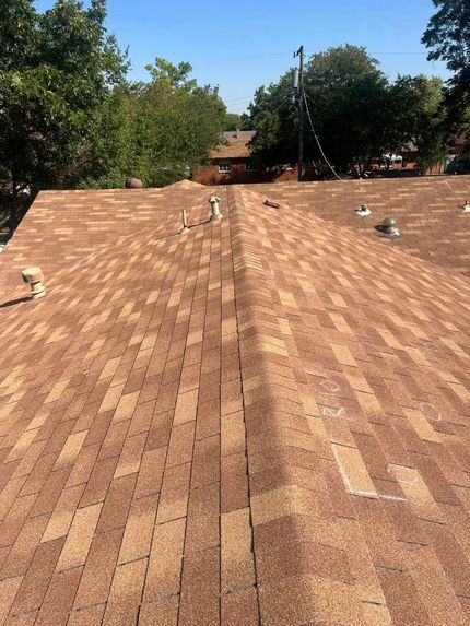 Rooftop with brown shingles and several roof vents, bordered by green trees under a clear sky