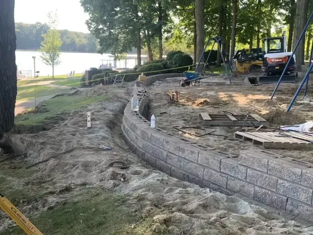 Construction of a curved stone retaining wall beside a lakeside yard with trees and tools.