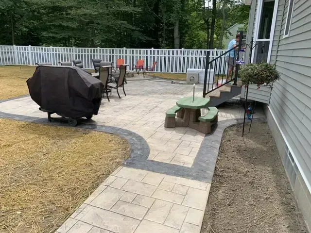 Backyard patio with paver walkway, covered grill, table and chairs, and steps to a house.