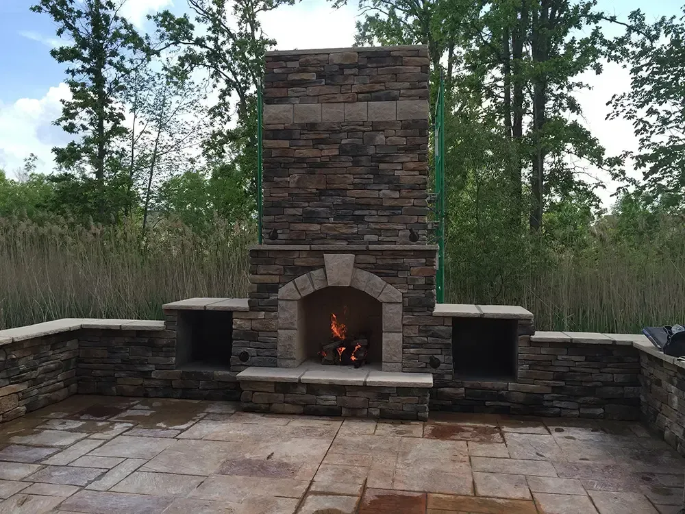 Stone outdoor fireplace with a small fire, surrounded by a patio and stone benches in a wooded setting.