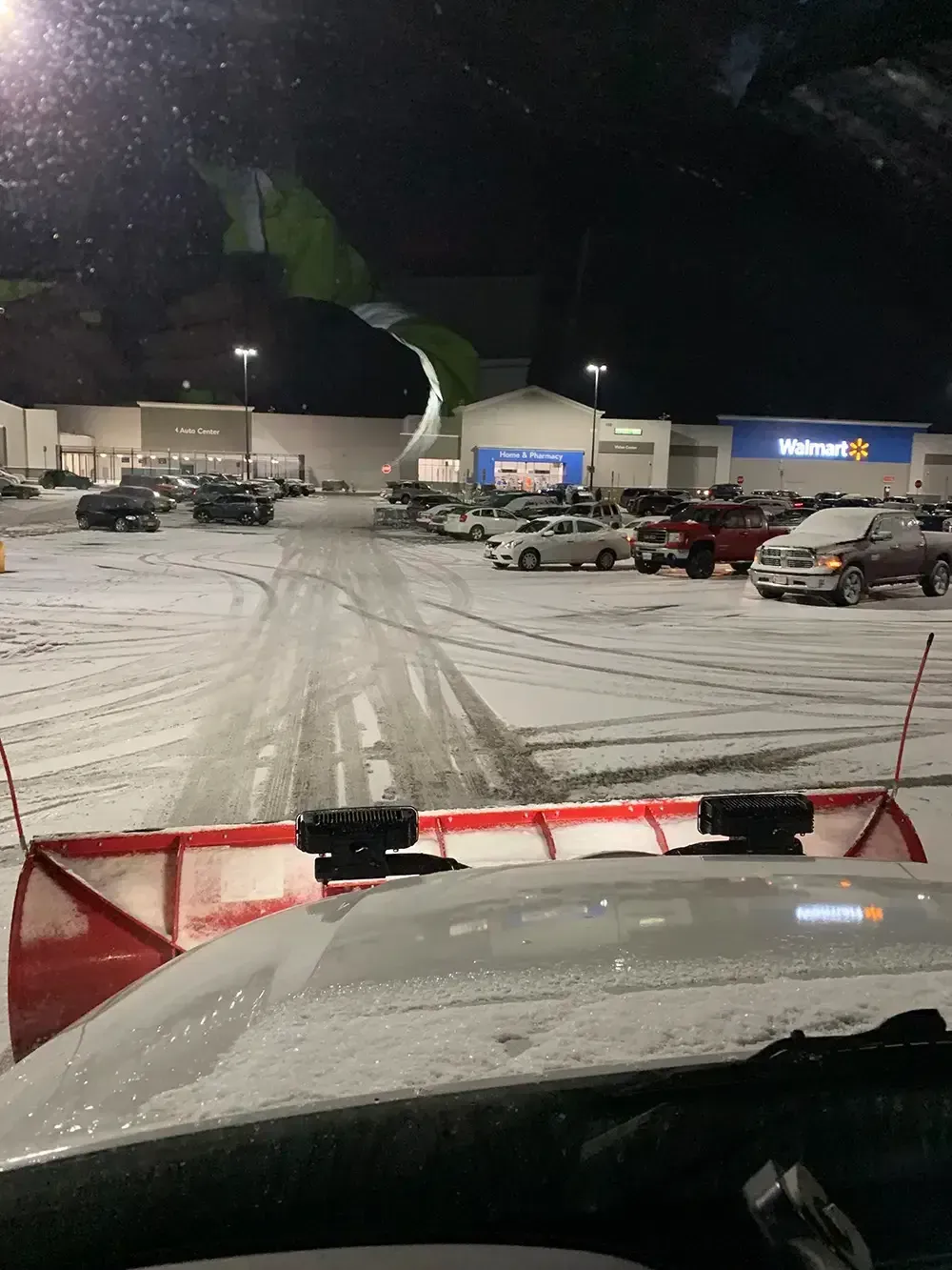 Snowy parking lot at night with plowed cars and storefront lights, viewed from inside a vehicle.