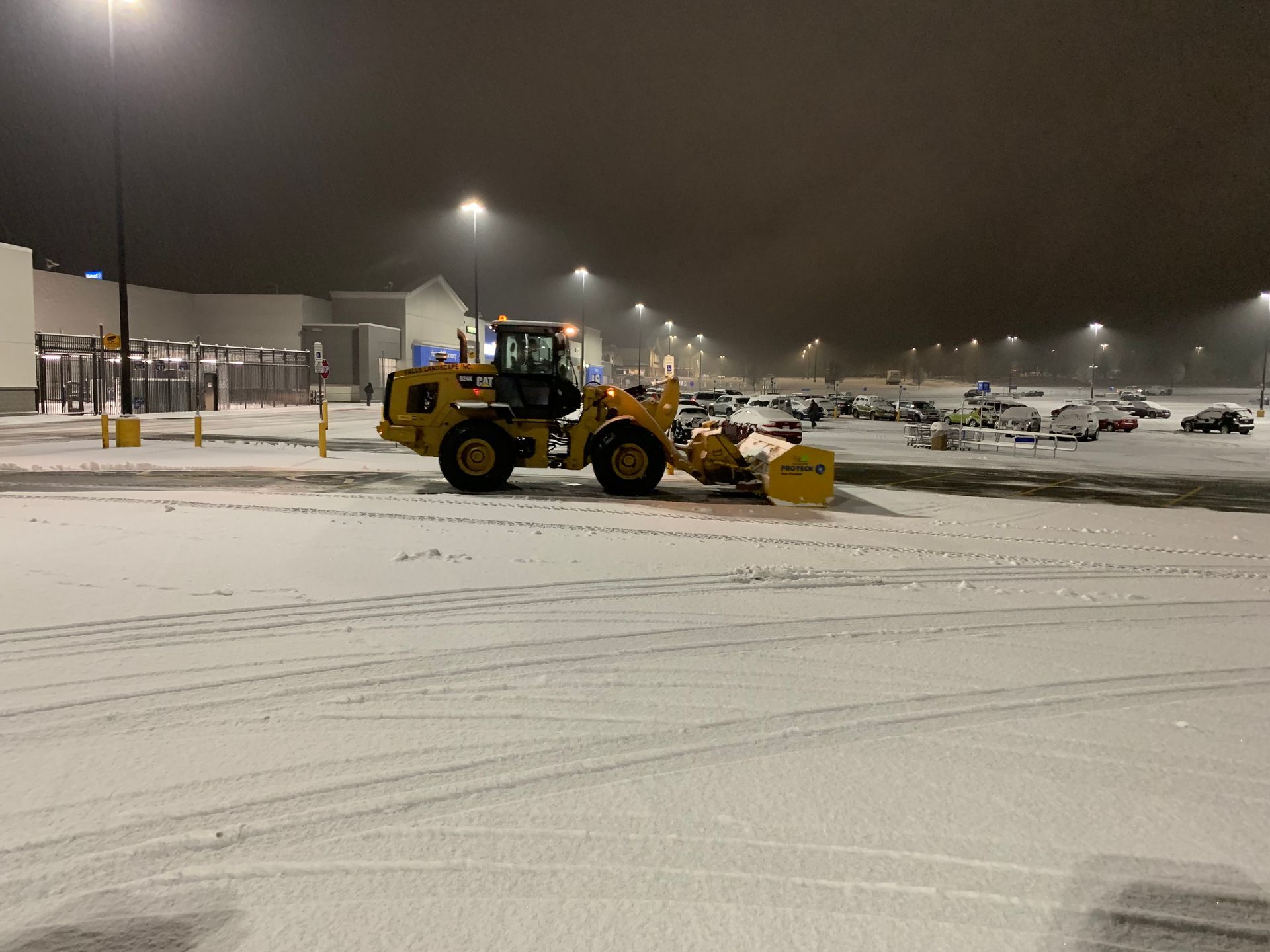 Yellow snowplow clearing a snowy parking lot at night under streetlights