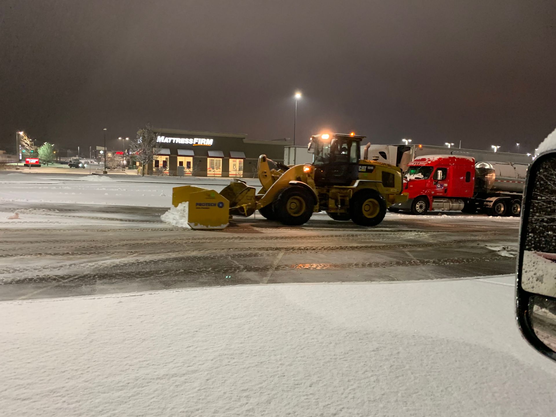 Snowy parking lot with a yellow front loader and red truck at night.