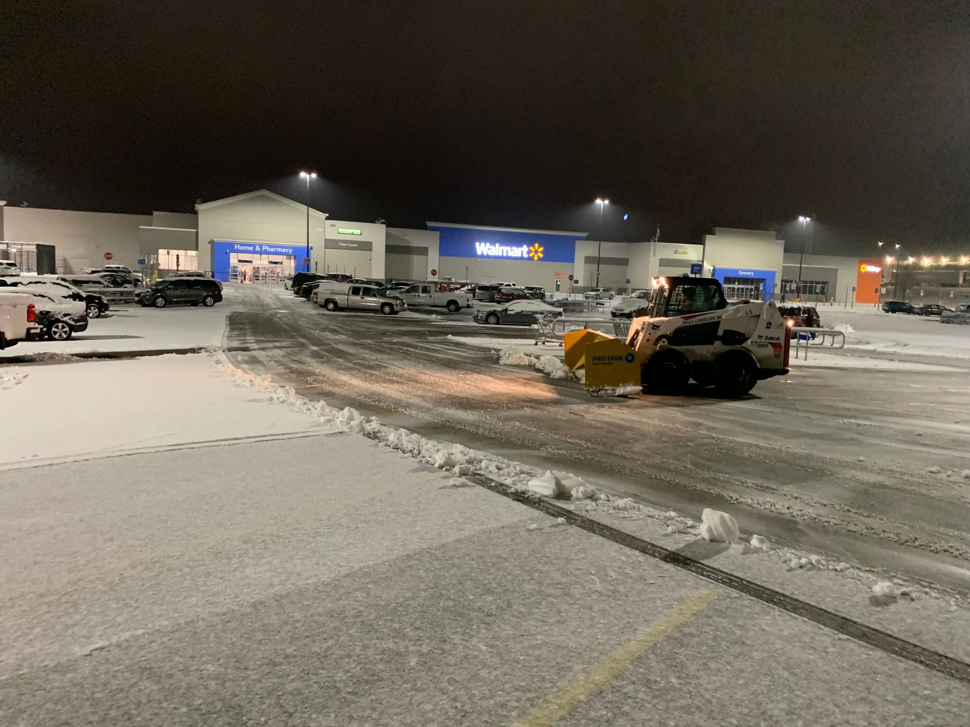 Snowy parking lot at night with plowed snow, parked cars, and bright store lights in the distance