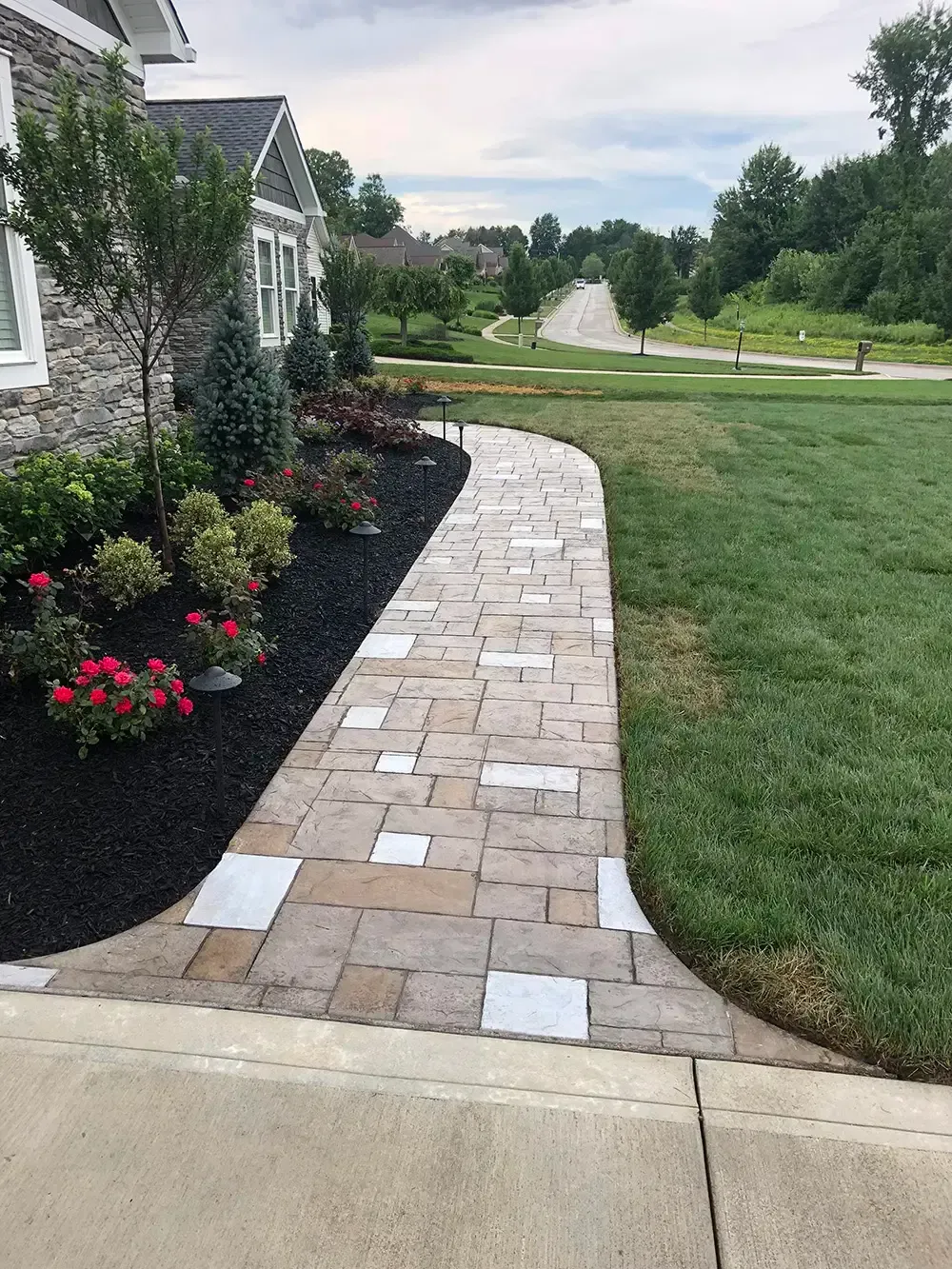 Curved brick walkway beside a stone house, bordered by flower beds and green lawn under cloudy sky
