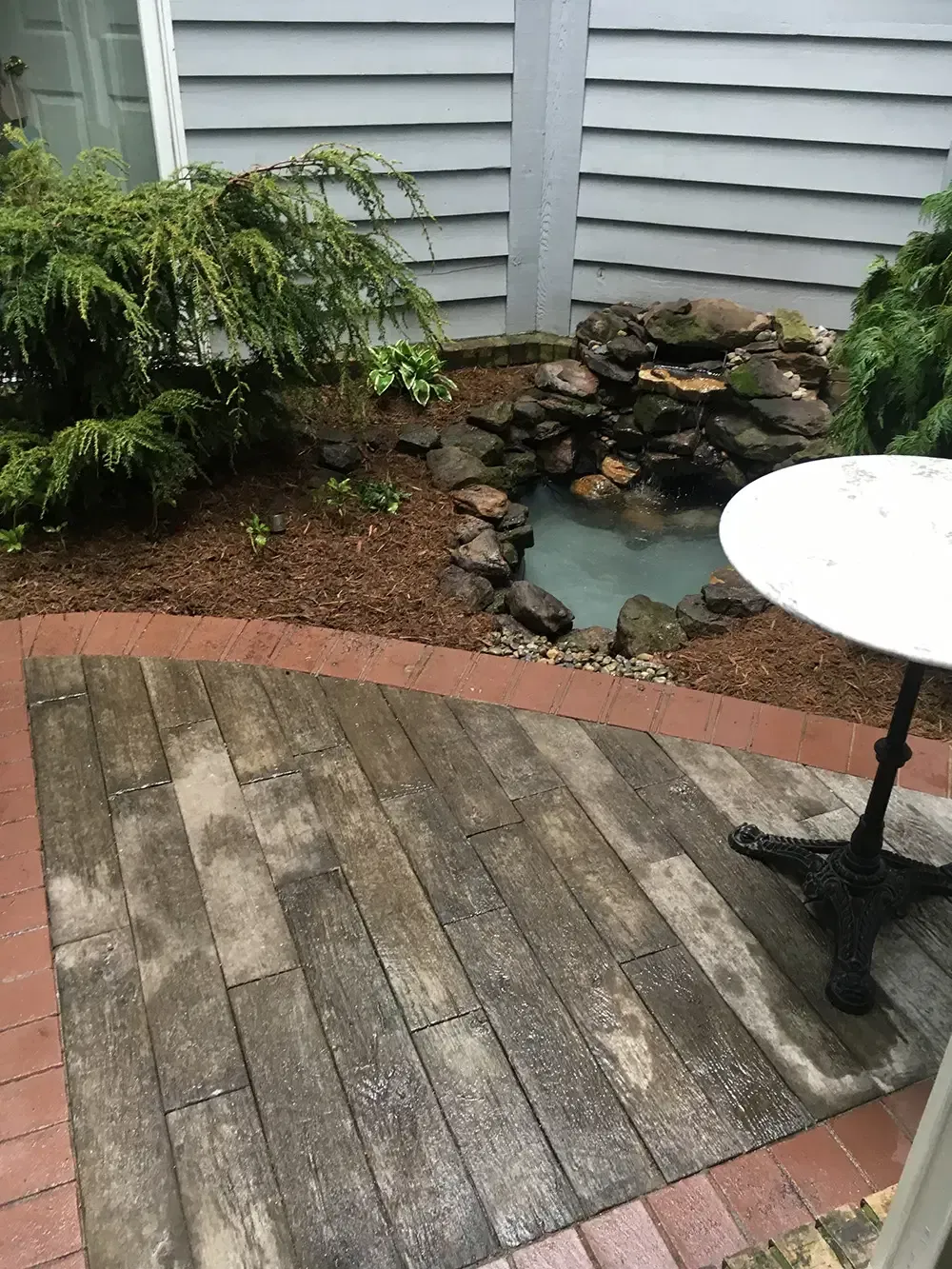Small patio with weathered wooden deck, brick border, and a rock garden pond beside white siding.