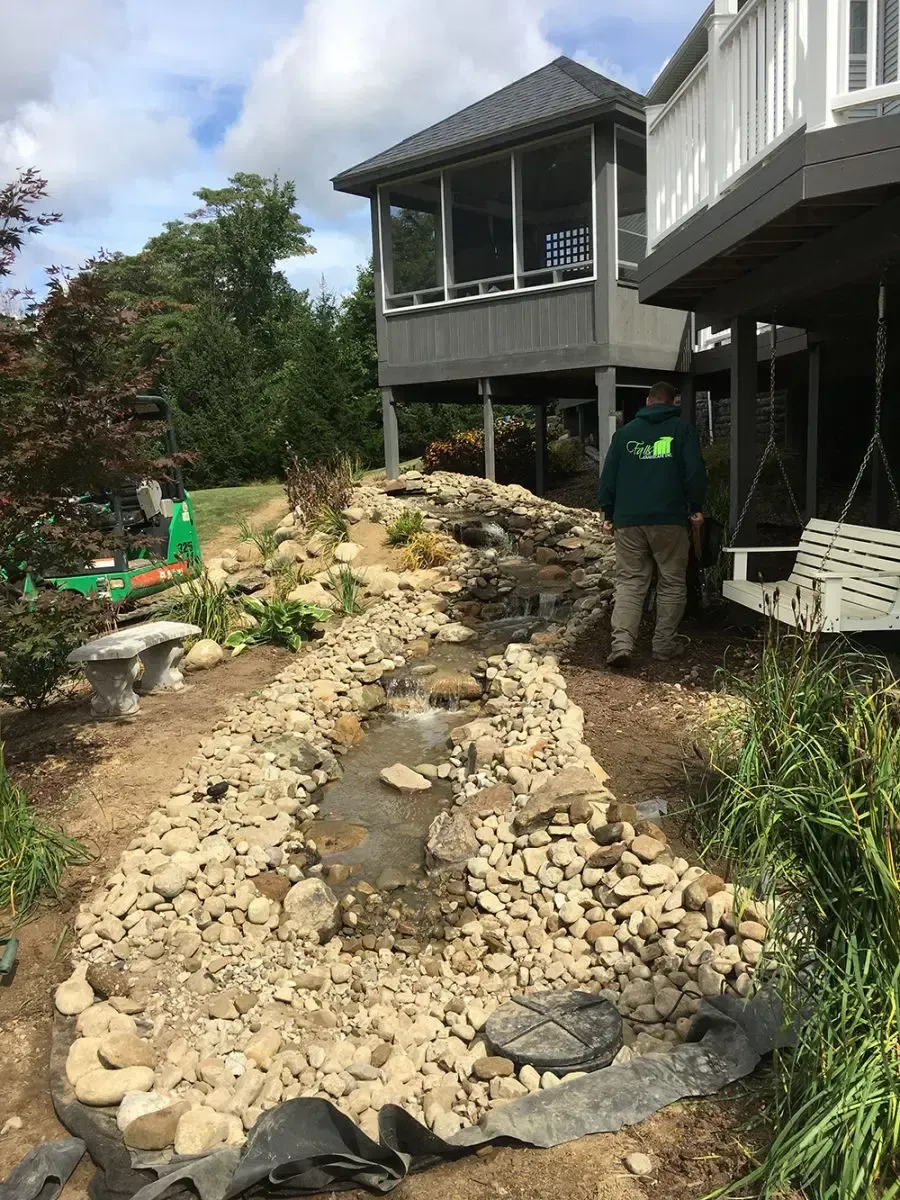 Workers beside a rocky drainage trench under a raised house in a yard