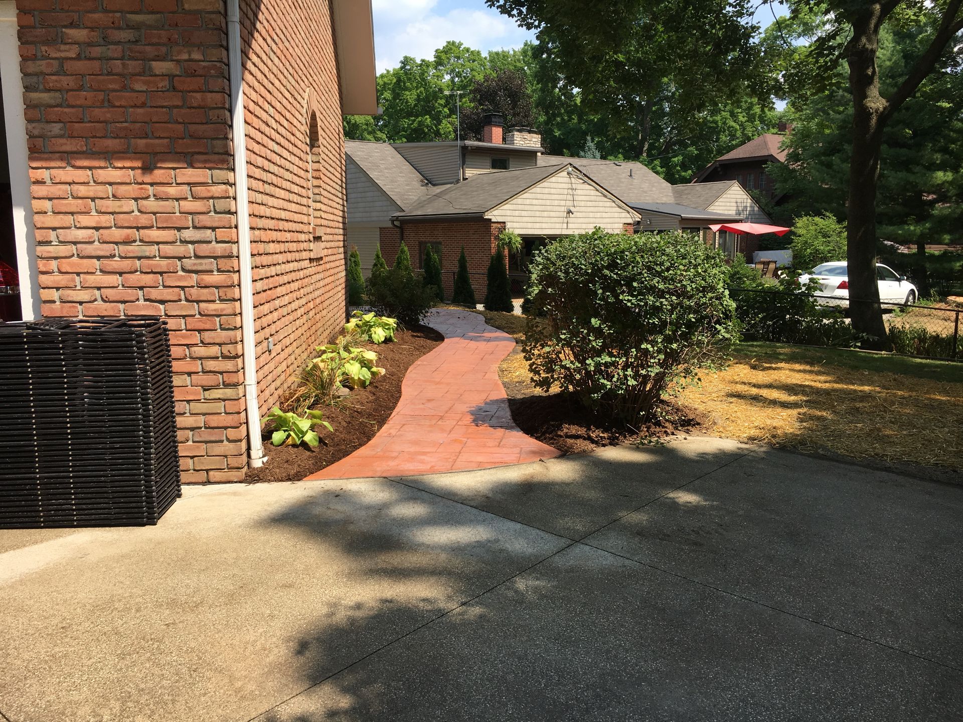 A brick walkway beside a brick building leads to a residential yard with bushes and parked cars beyond.