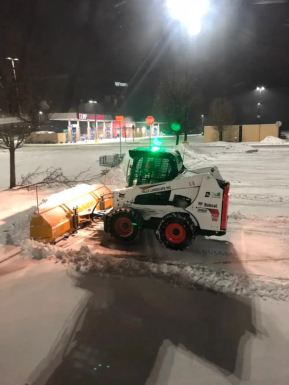 Snowplow clearing a snowy parking lot at night under bright lights, with green beacon flashing.