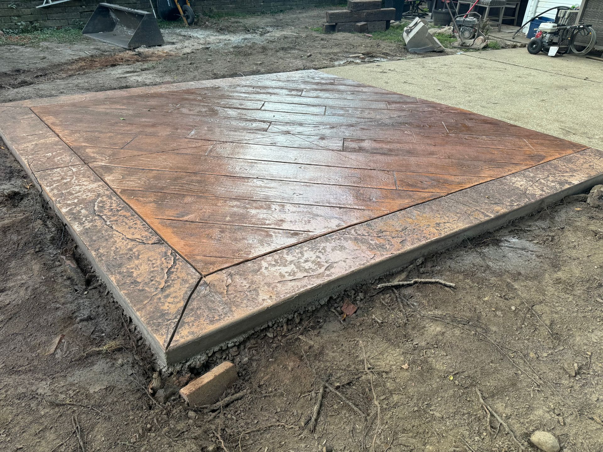 Rust-colored stamped concrete slab or patio in an outdoor dirt yard, shown at a slight angle.