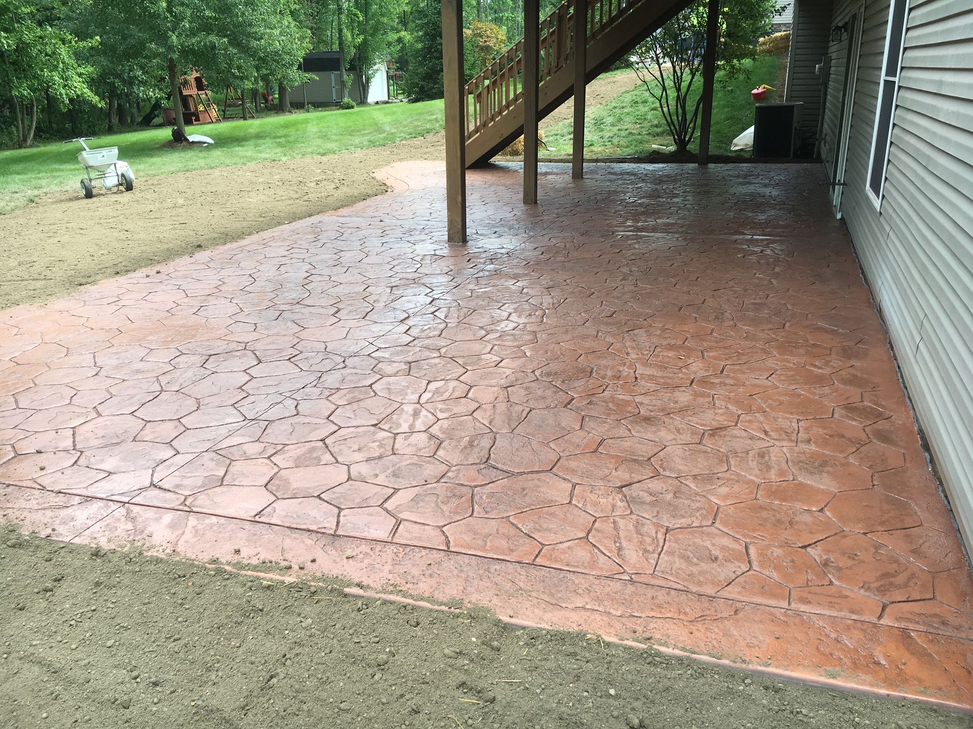 Covered red stamped concrete patio beside a house, with stairs, siding, and a grassy yard in the background