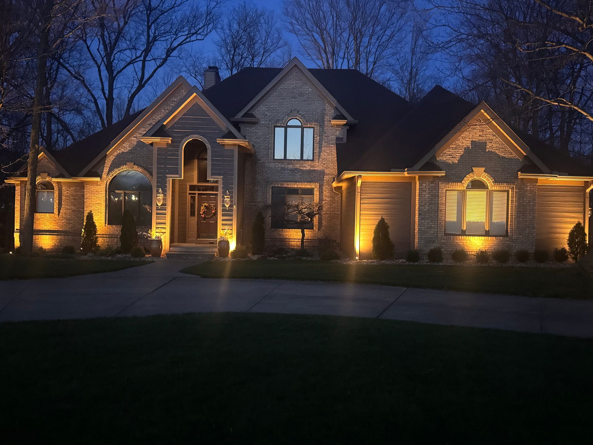 Large brick suburban house at dusk, warmly lit with exterior lights and dark trees silhouetted against the sky