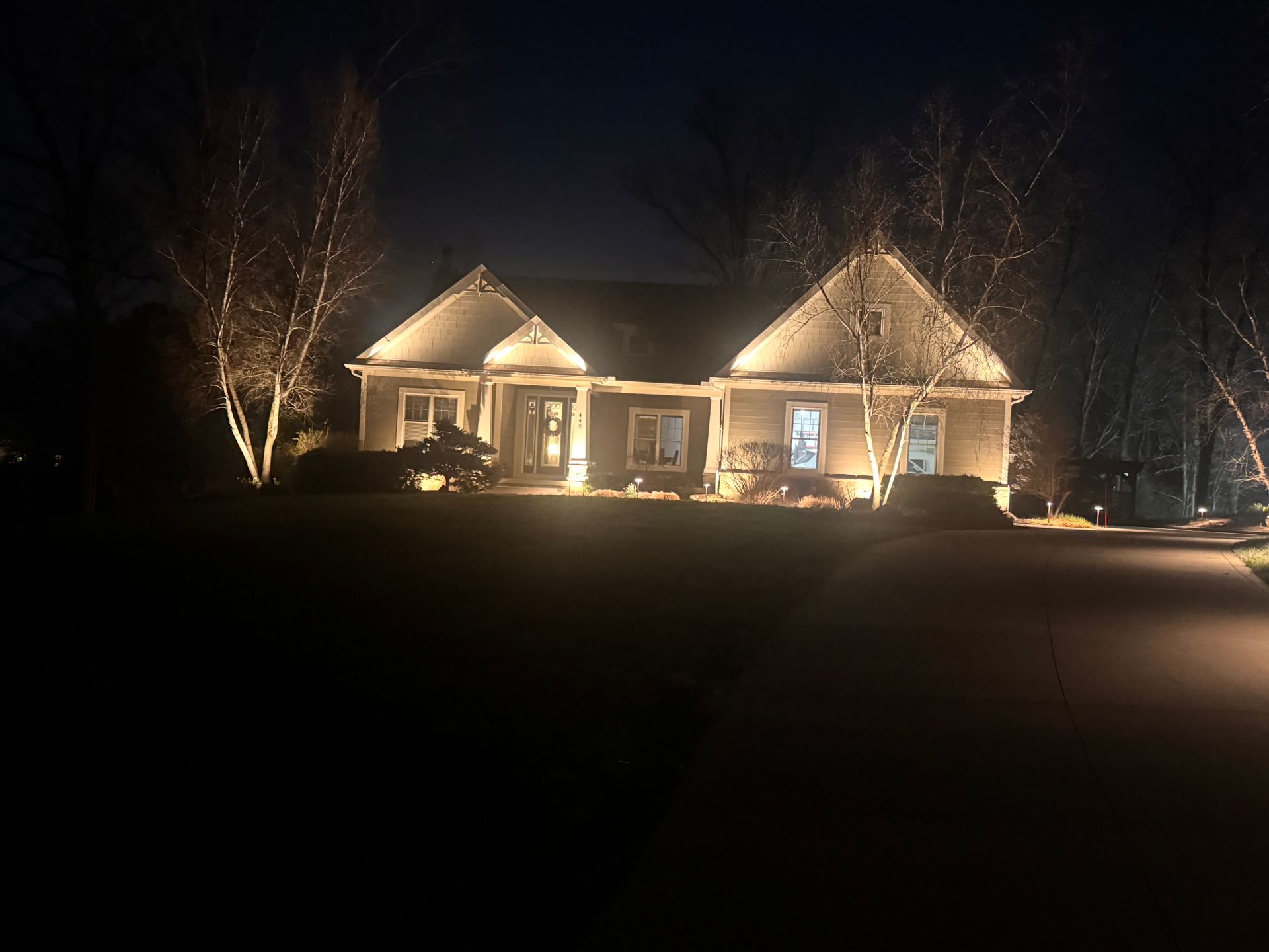 Single-story house lit by warm lights at night, with illuminated trees and a dark foreground