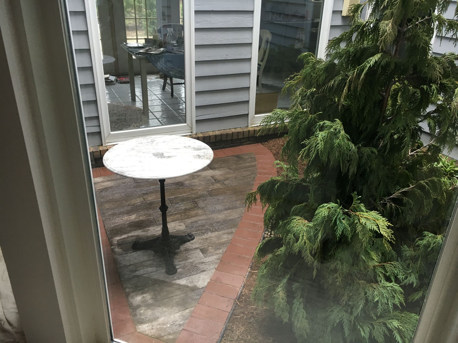 Small patio with a white round table, brick walkway, and a green potted tree beside a house siding wall.