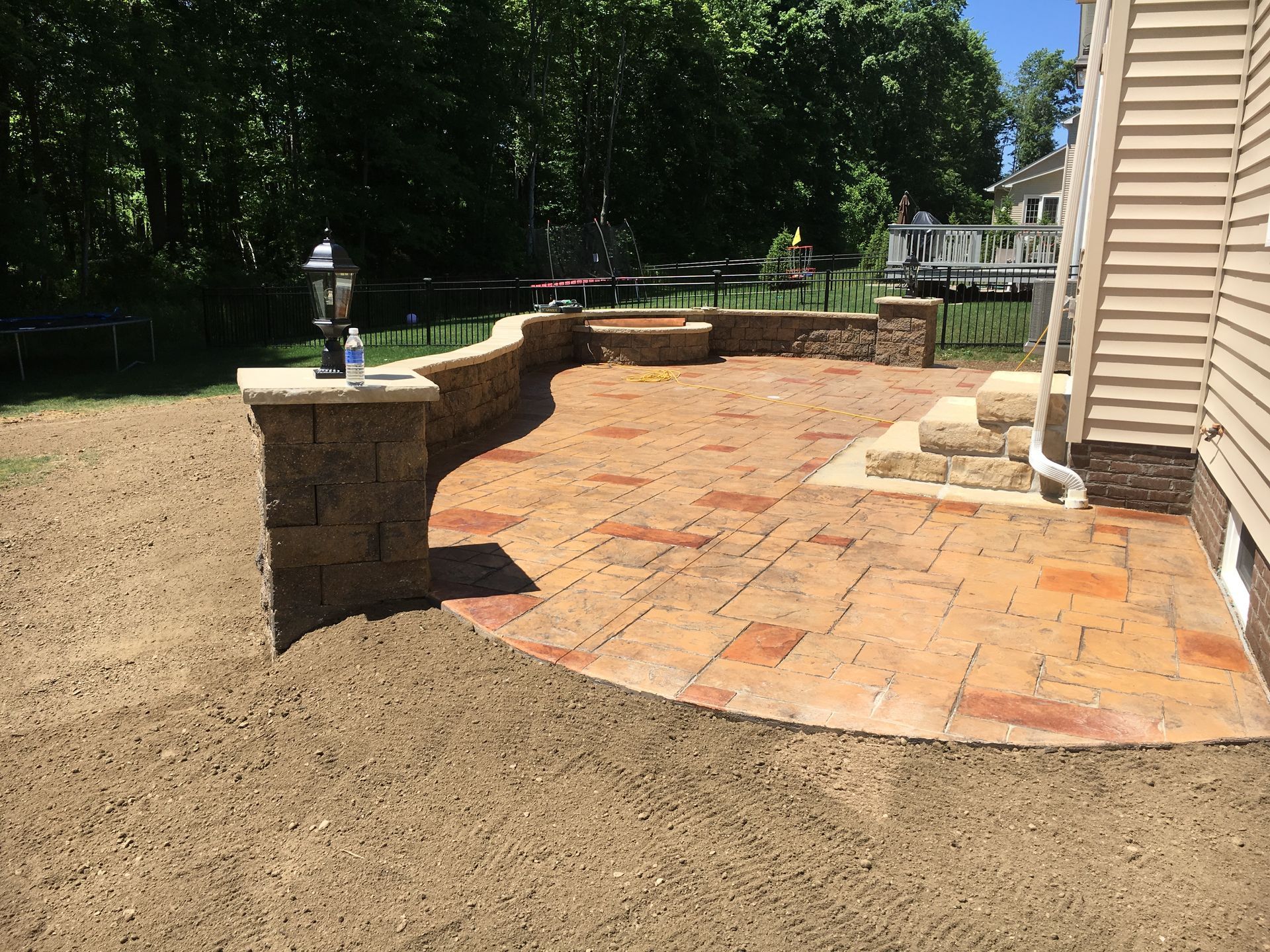 Curved brick patio with low stone wall beside a beige house, bordered by gravel and trees