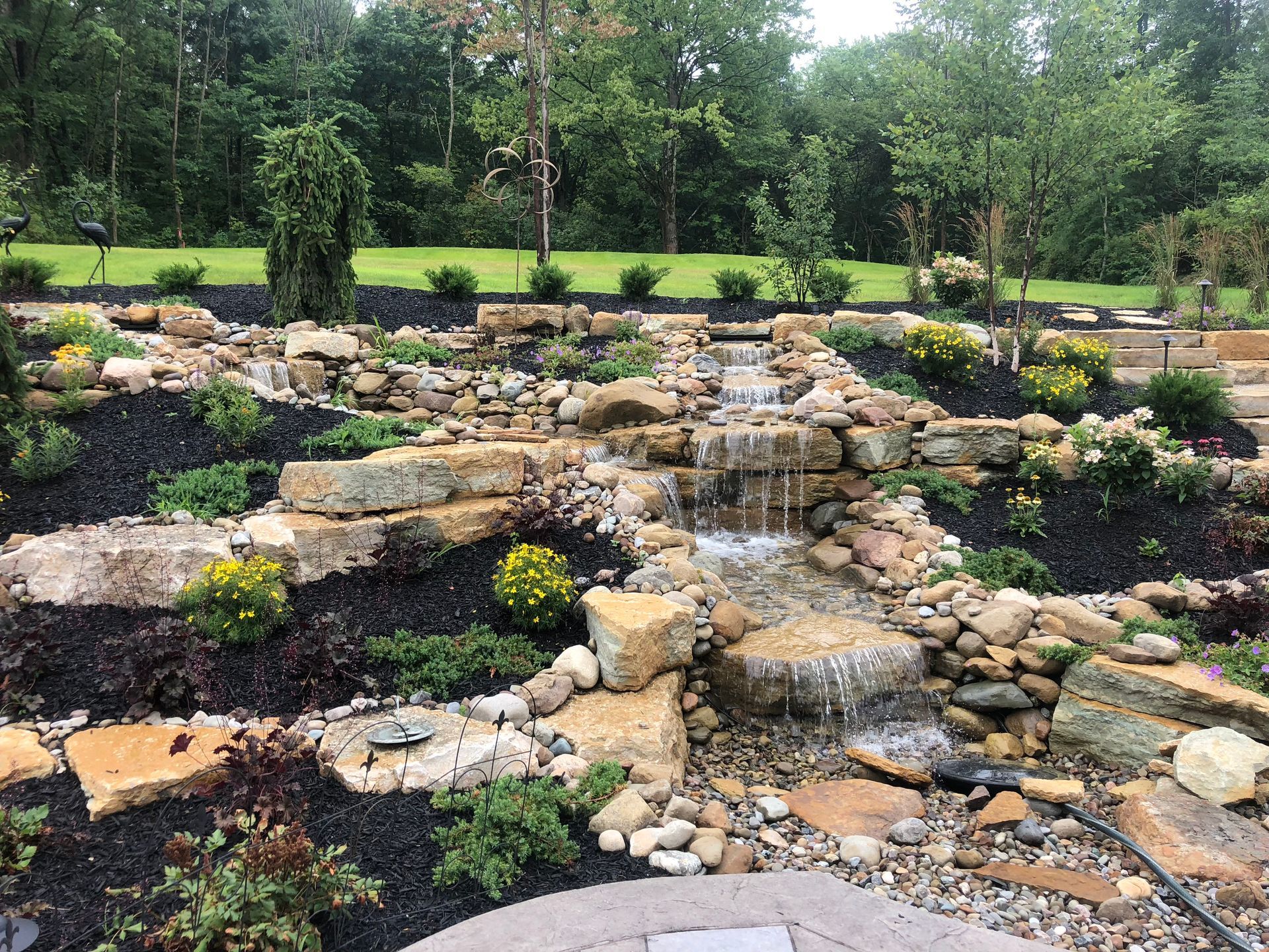 Stone waterfall feature in a landscaped garden with rocks, plants, and trees in the background