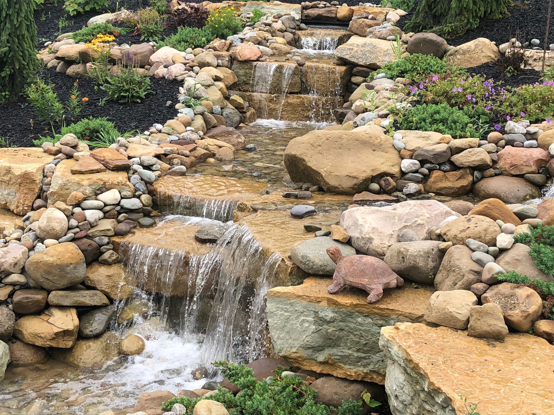 Stone waterfall in a landscaped garden with a small dog standing on rocks near the water