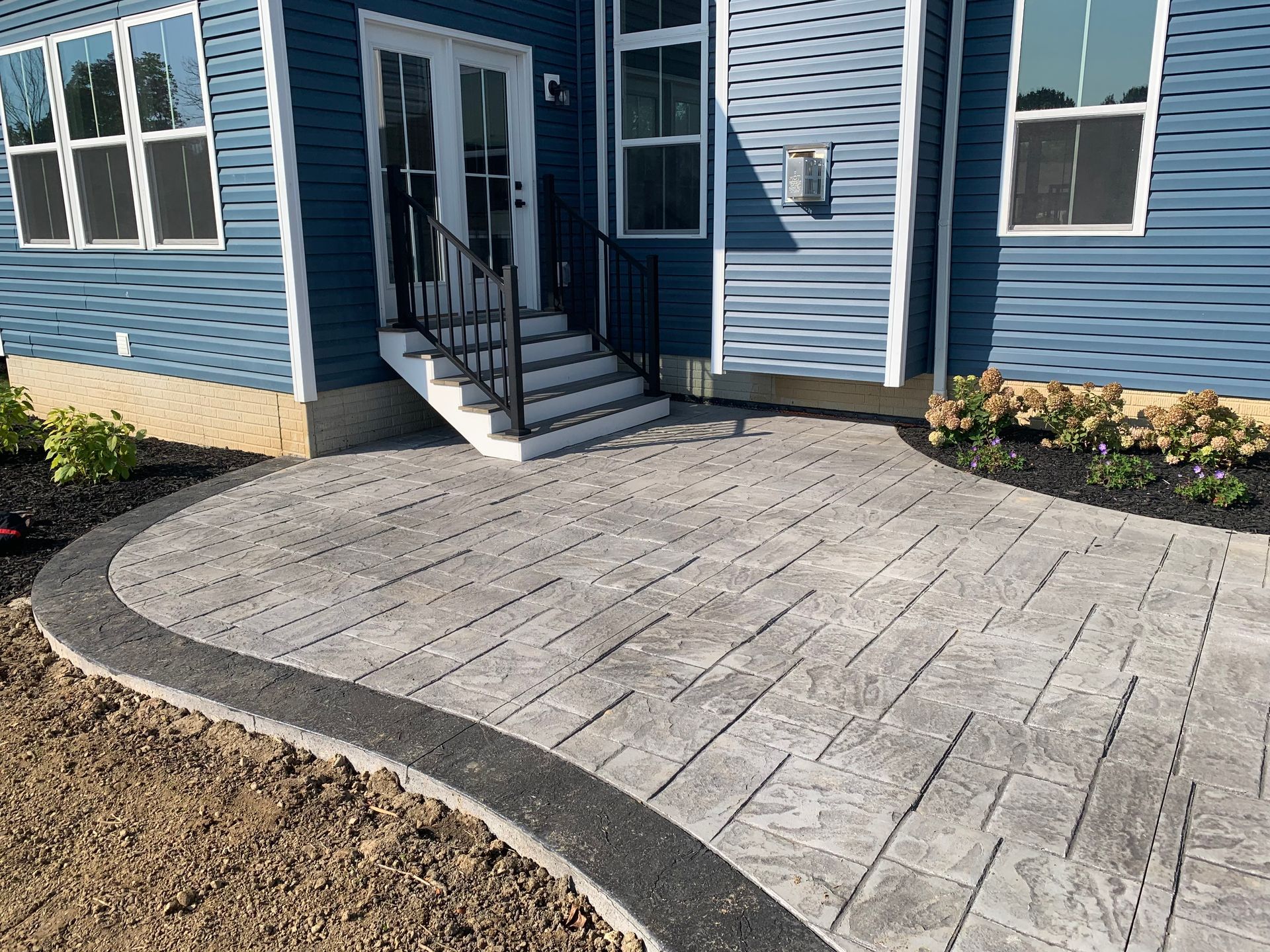 Curved stone patio walkway leading to a blue house entrance with steps and flower beds