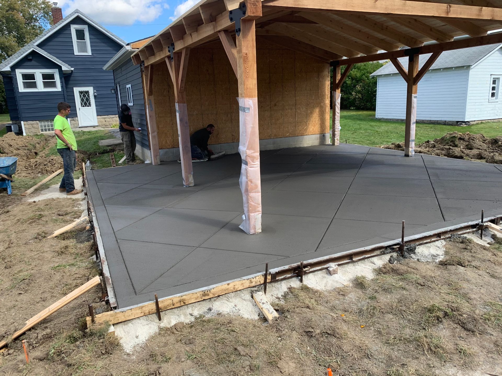 Concrete slab being poured for a covered patio beside a house, with workers and wooden support posts.