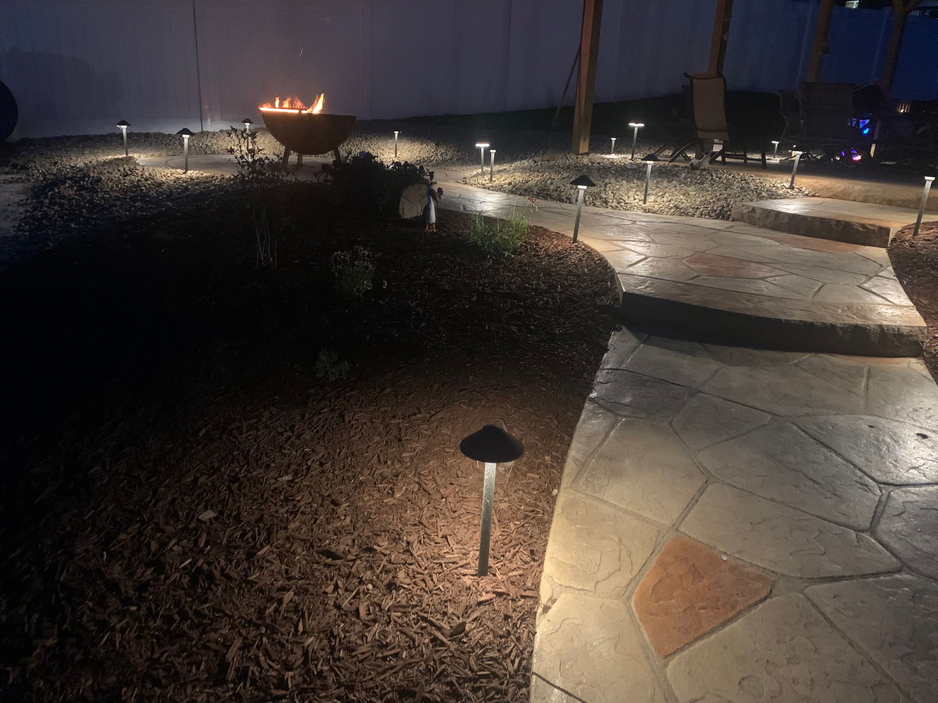 Nighttime stone path with small garden lights, gravel beds, and a lit fire bowl in a landscaped yard.