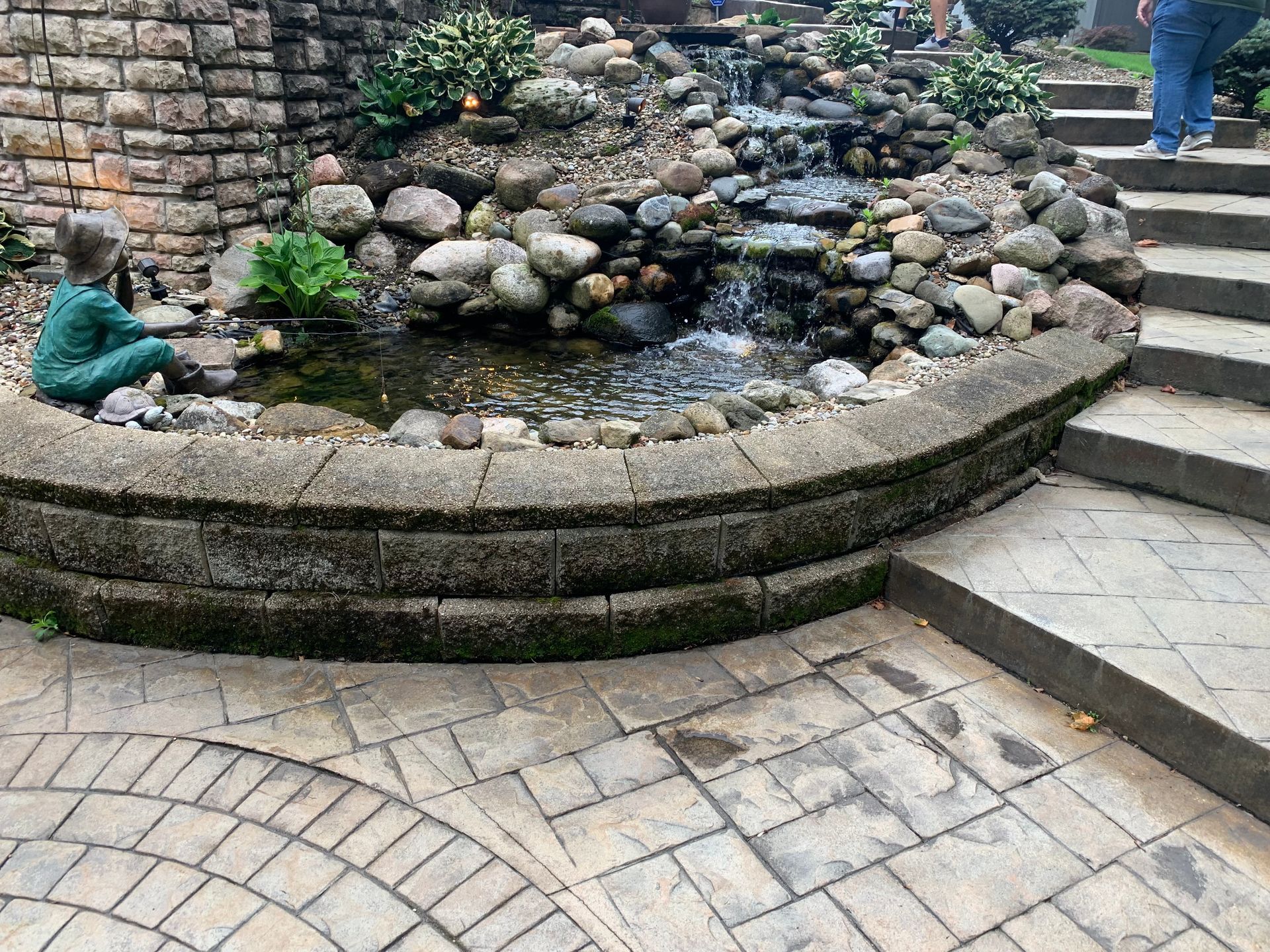 Stone pond with rocks and plants beside curved brick steps and a paved patio