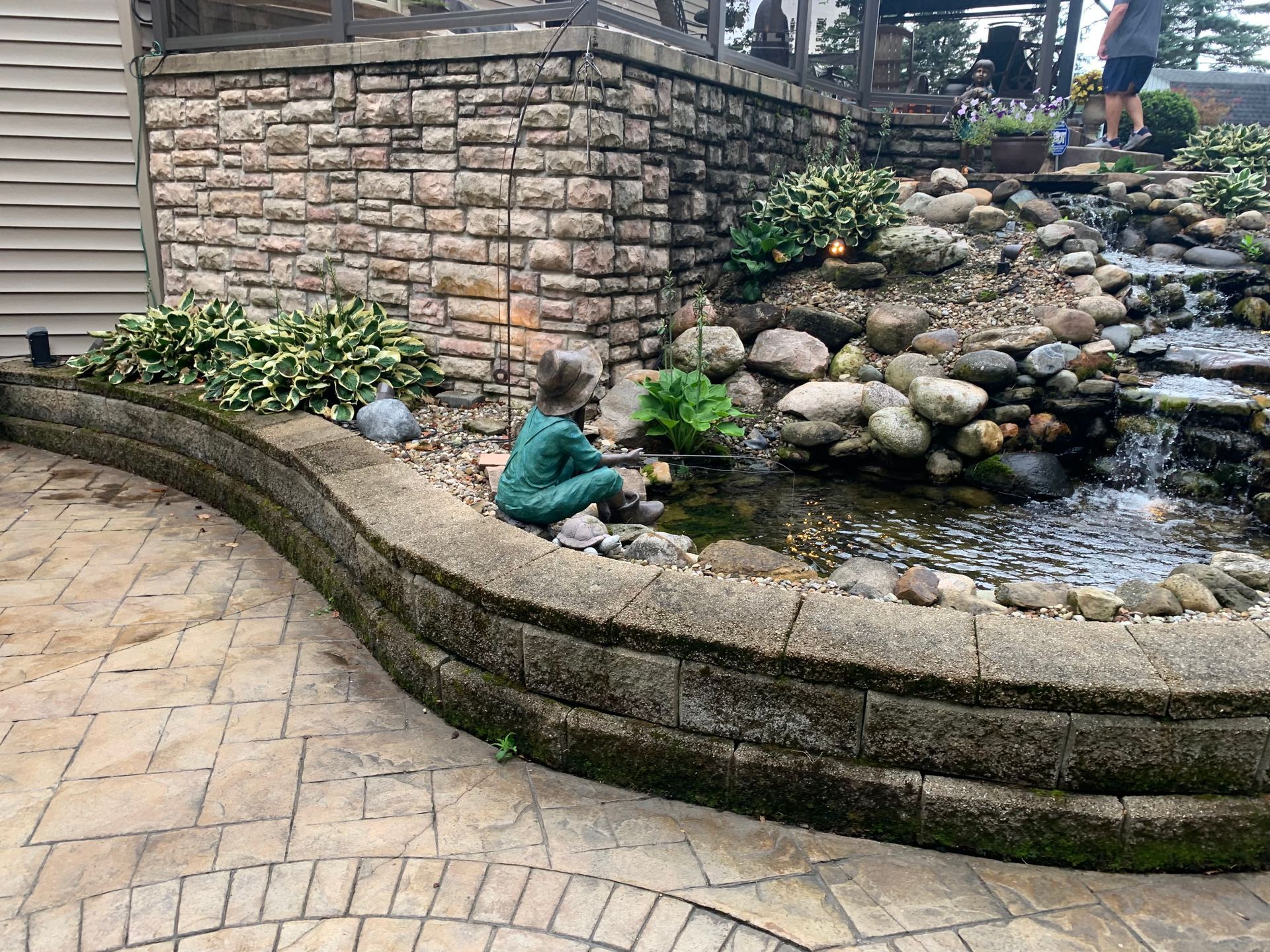 Child sitting by a backyard rock pond and waterfall beside a stone wall and curved patio