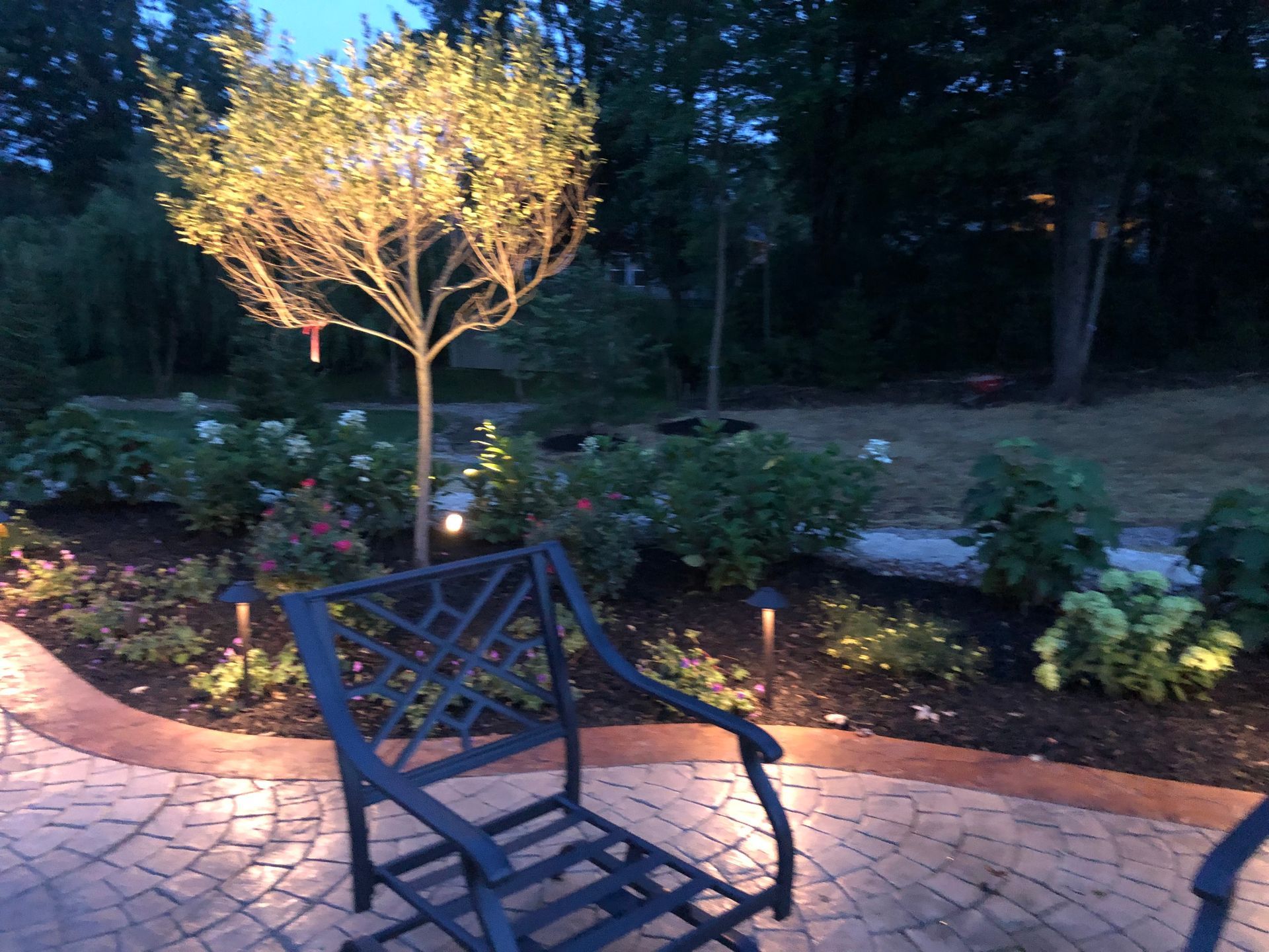 Lit garden patio with glowing tree, flower beds, and a black metal chair at dusk