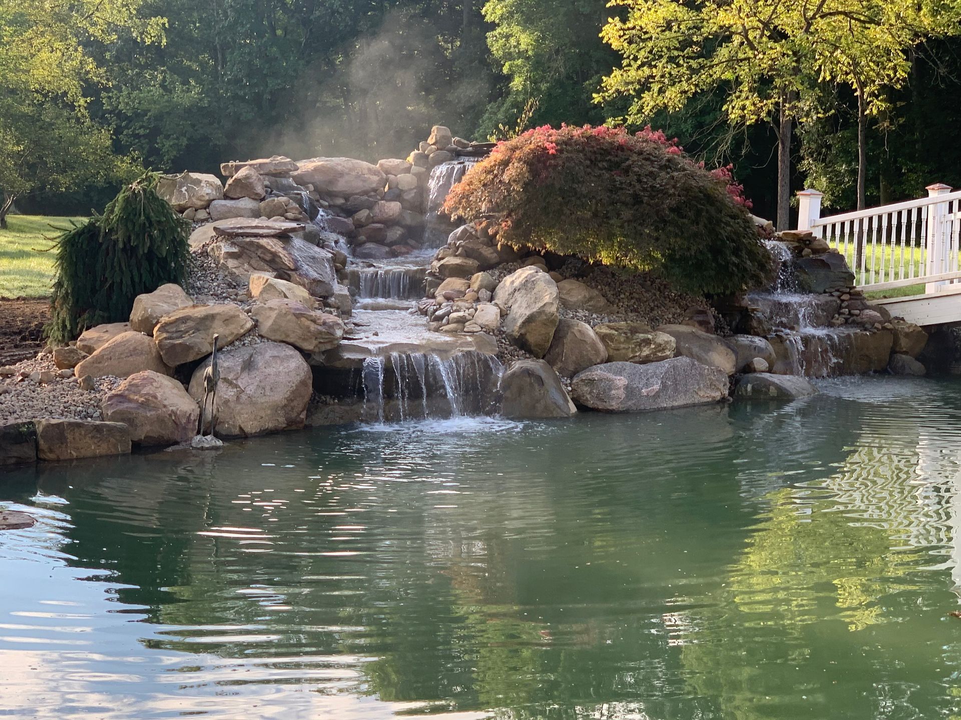 Rock waterfall cascading into a calm pond, surrounded by boulders, greenery, and a white fence.