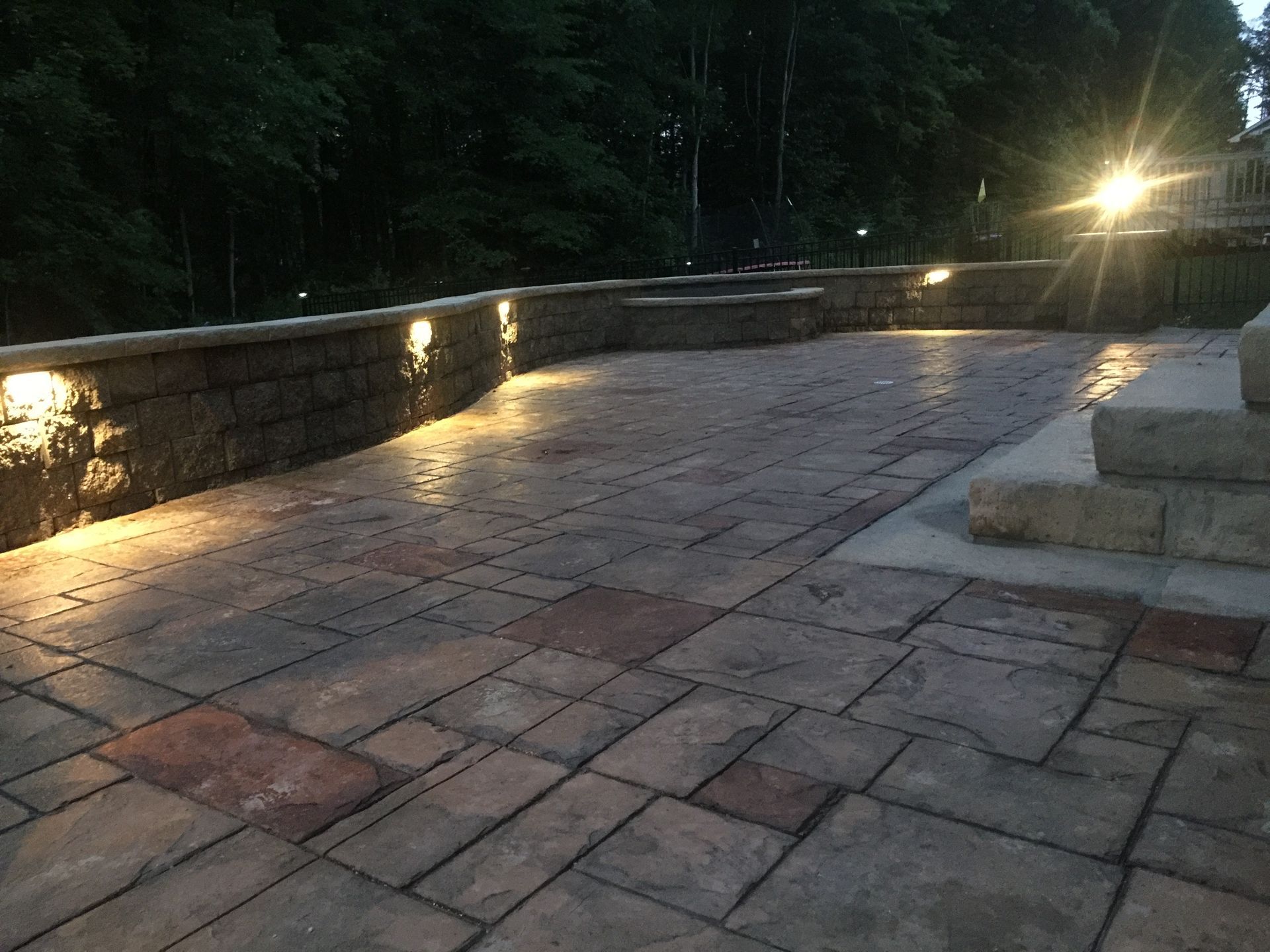 Stone patio walkway beside a low-lit wall at dusk, leading into a dark wooded area