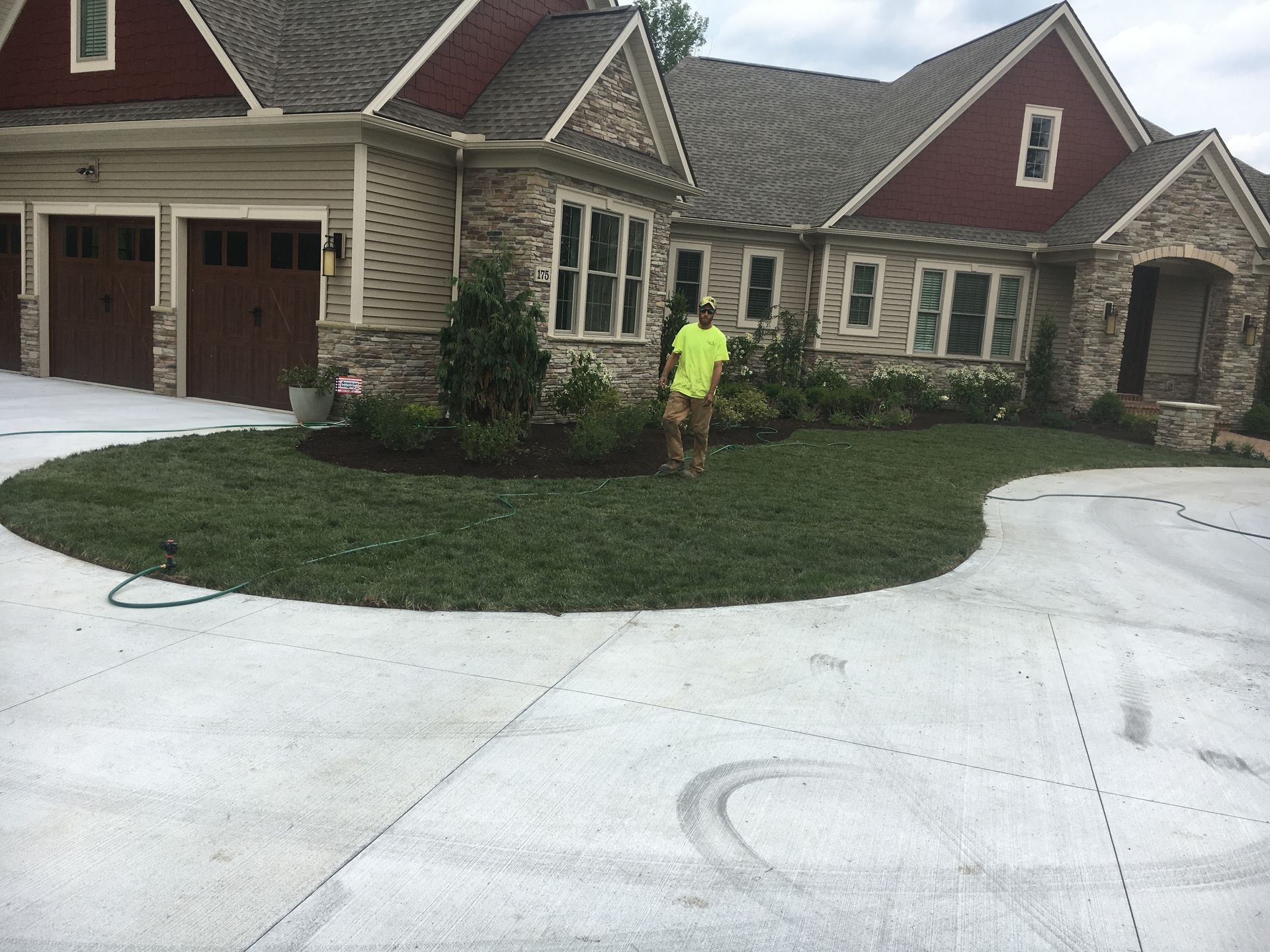 A person in a neon shirt working on a front lawn beside a large brick house and curved driveway.