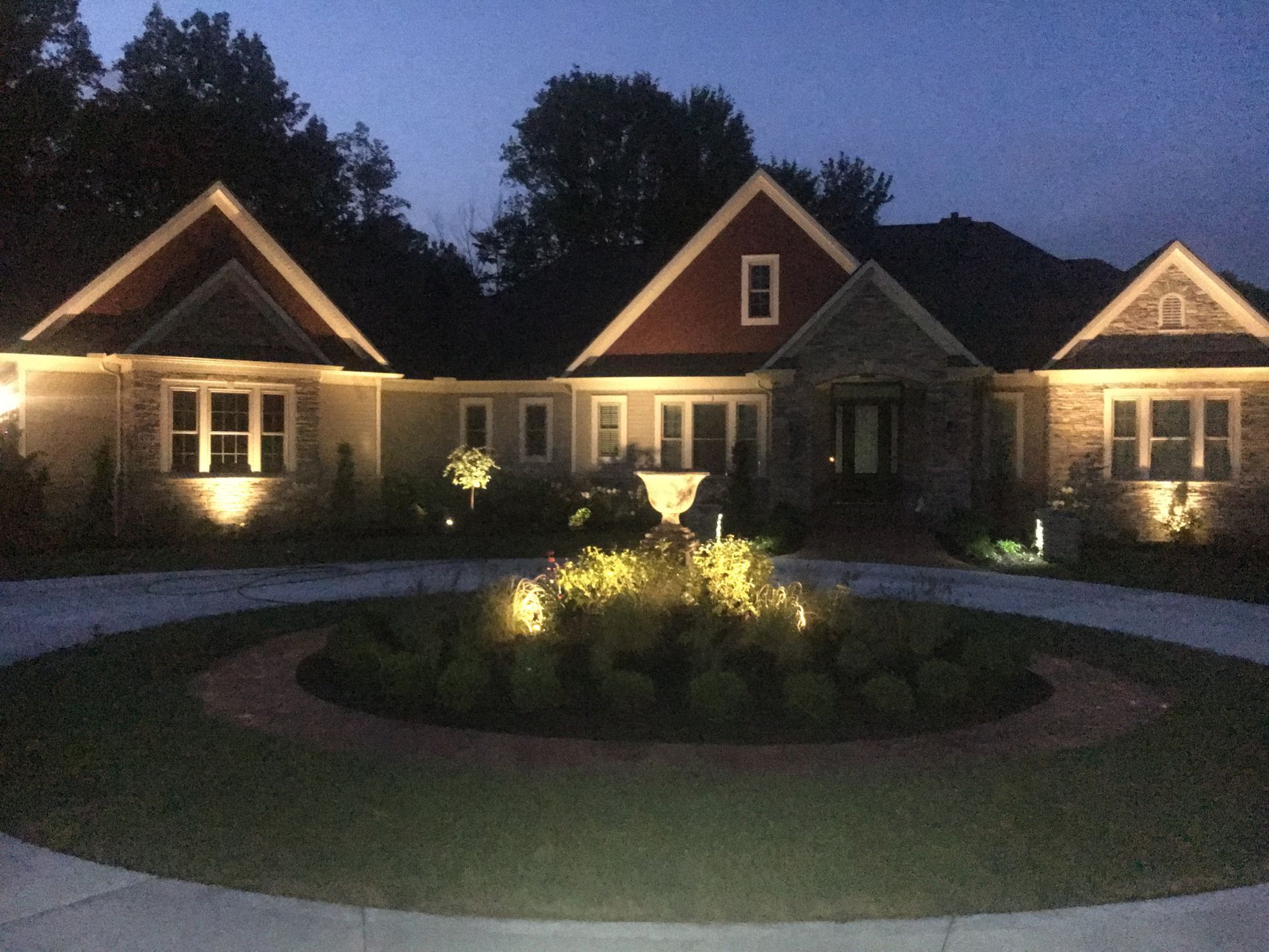 Illuminated house at dusk with peaked roofs, landscaped lawn, and circular driveway