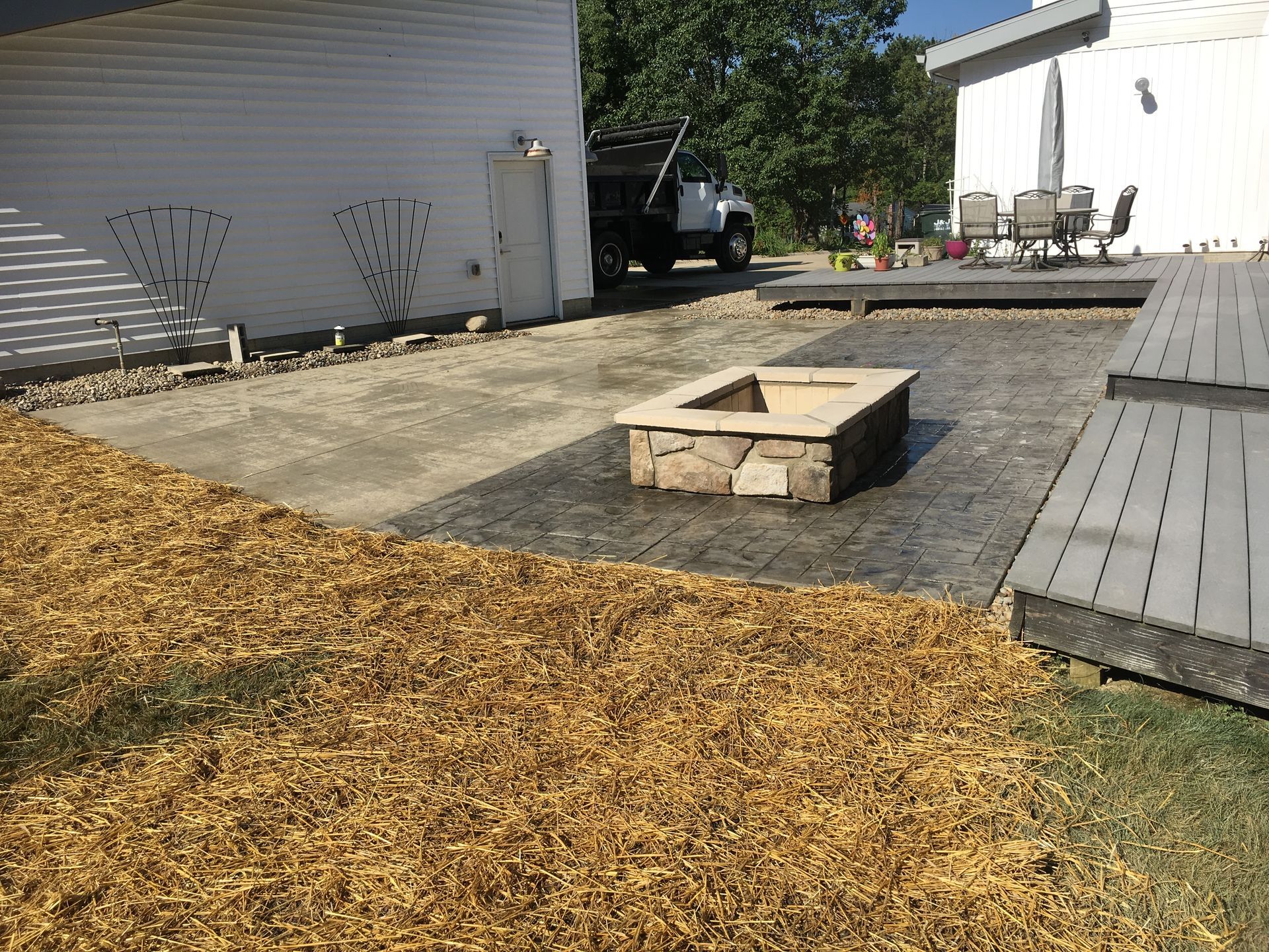 Backyard with a square fire pit, wood chips, paved patio, and two buildings under a clear sky.
