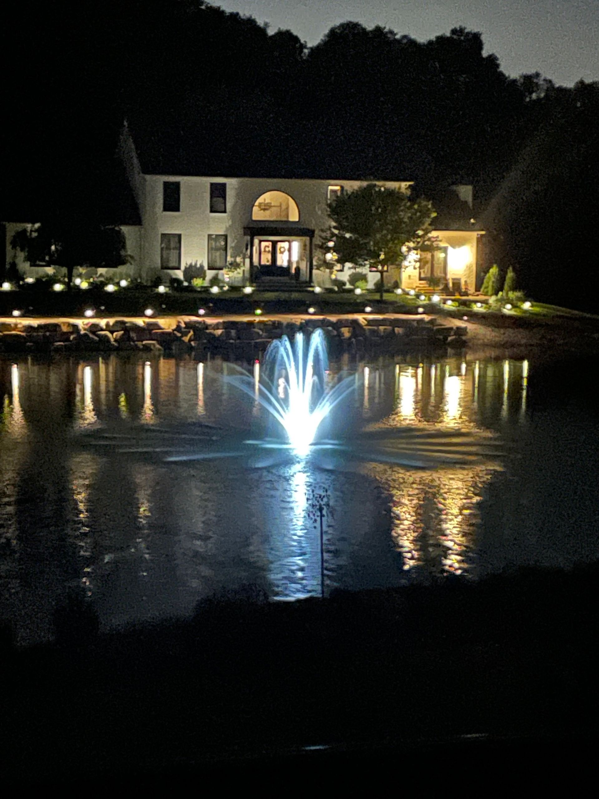 Nighttime view of a lit house and fountain reflecting in a calm pond