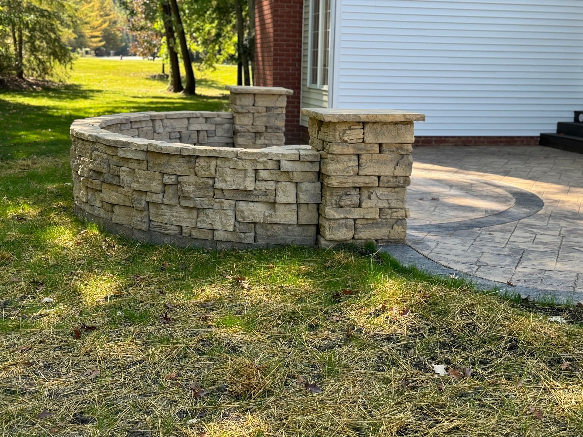 Curved stone patio wall beside a house, with a paved walkway and grassy yard in sunlight