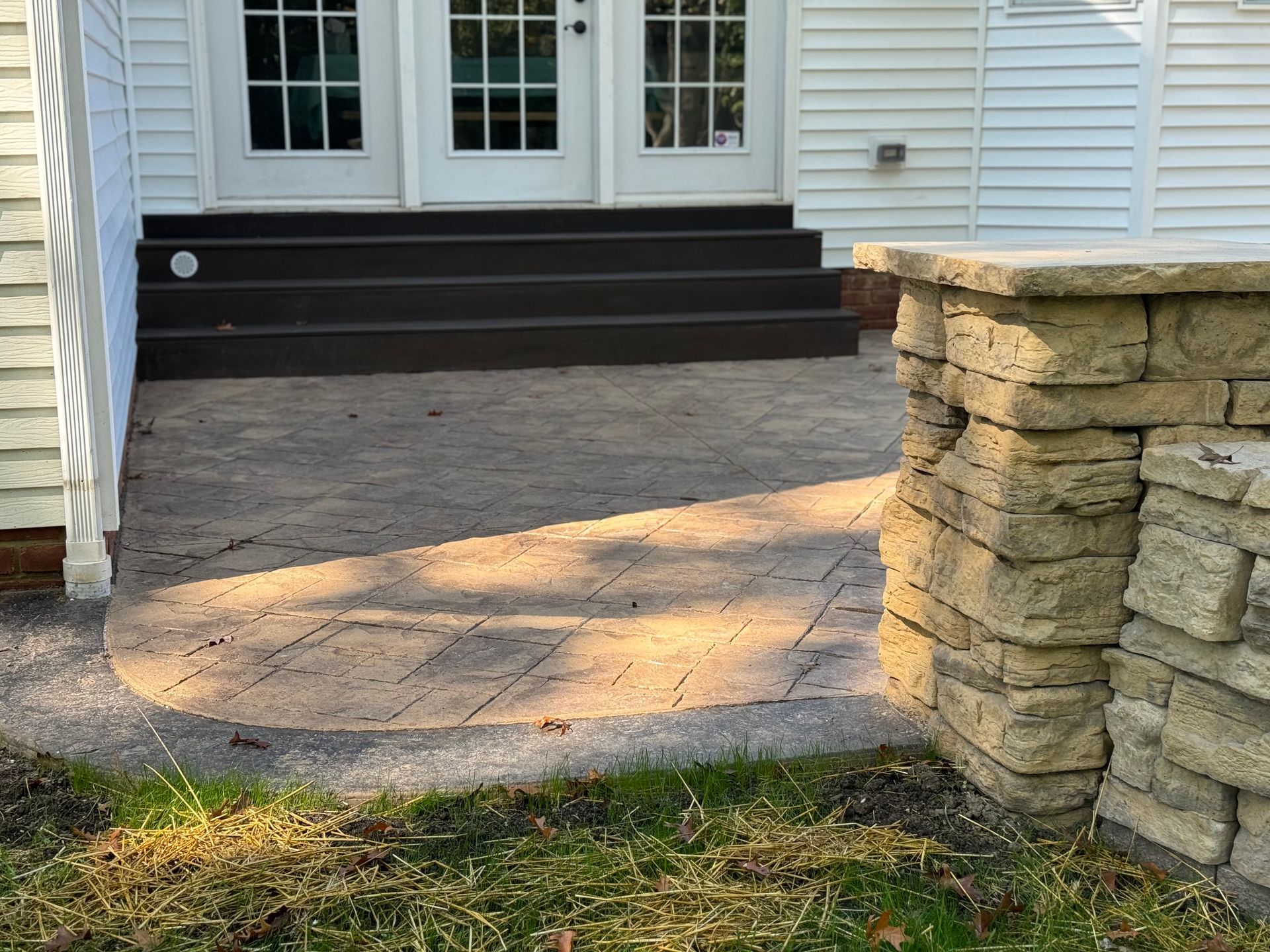 Concrete patio with dark steps leading to white French doors beside a stone wall.