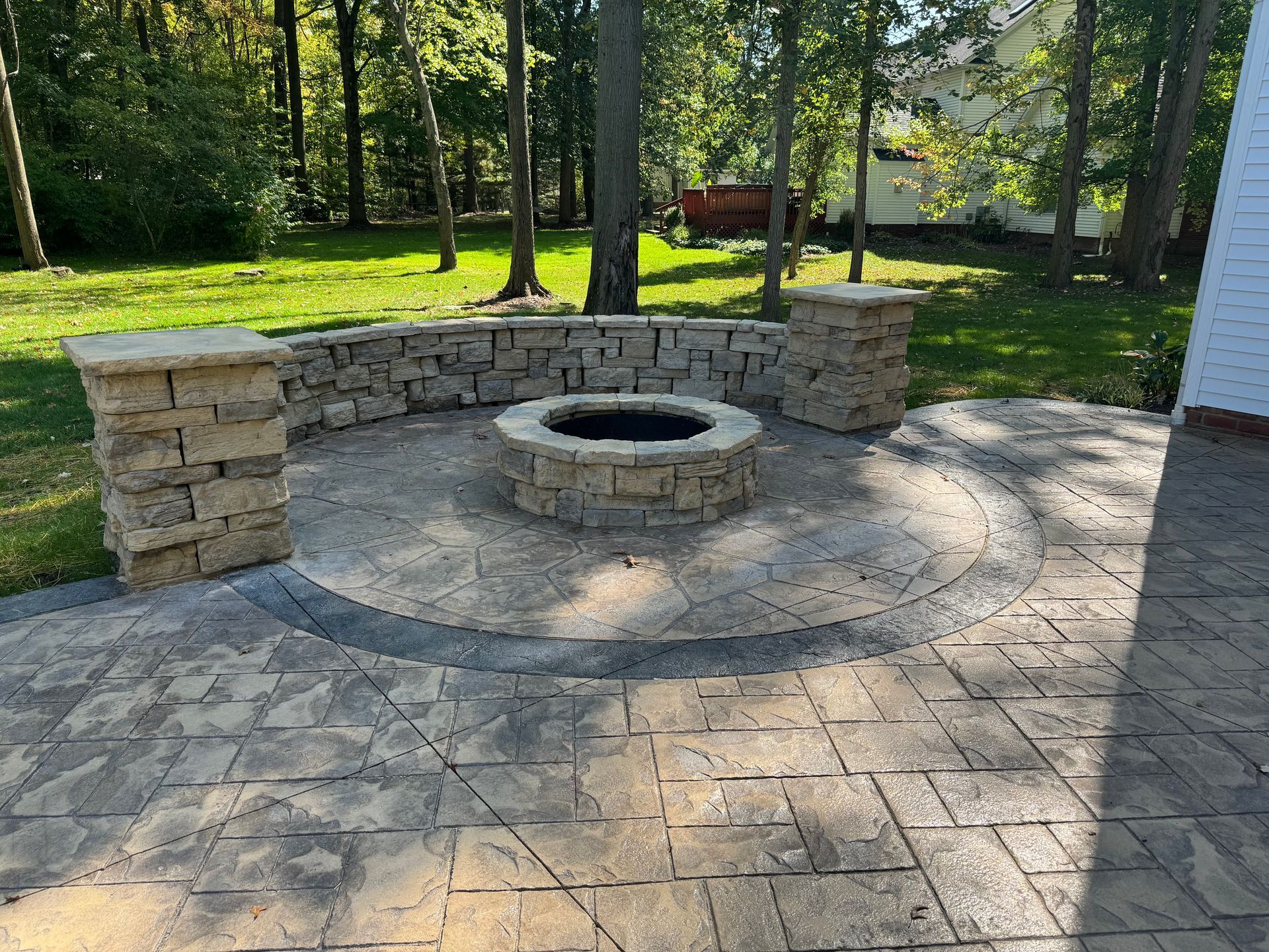 Stone patio with curved benches and a circular fire pit in a wooded backyard