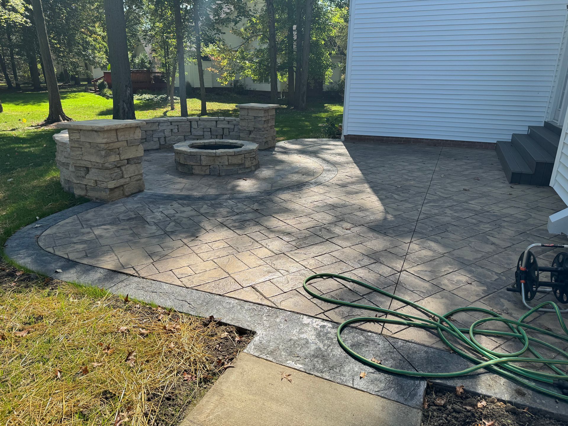 Stone patio with fire pit, bordered by grass and trees beside a white house wall.