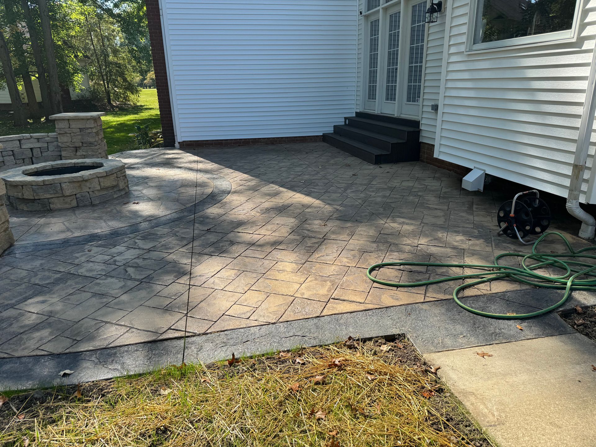 Backyard stone patio with a fire pit, garden hose, and white house siding in sunlight
