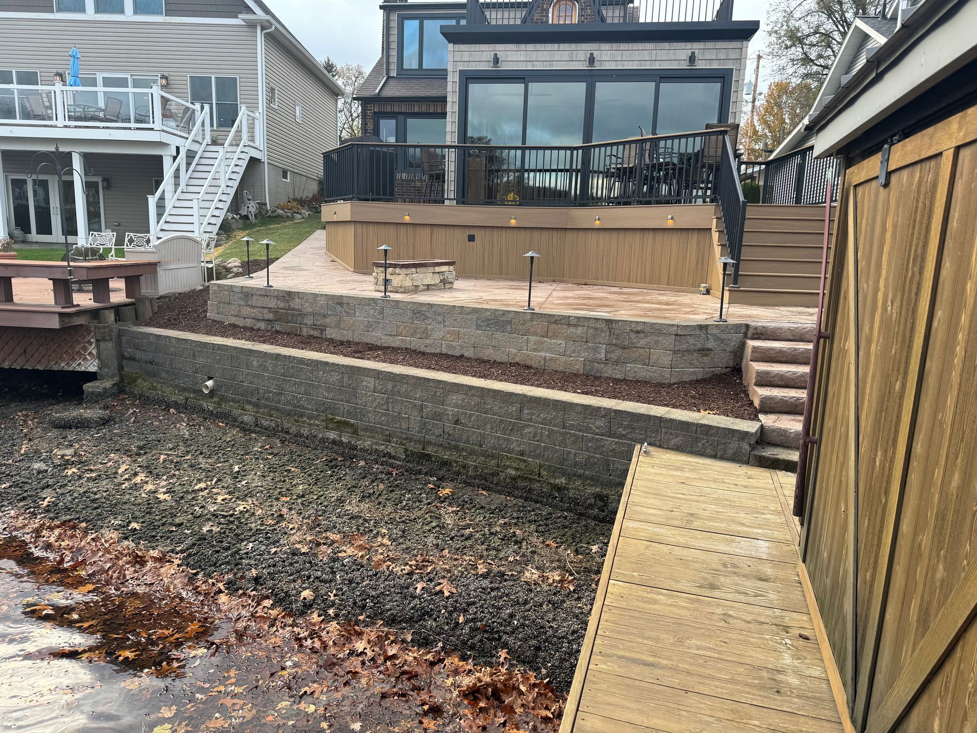 Backyard with a raised wooden deck, stone retaining wall, and narrow dock over water.
