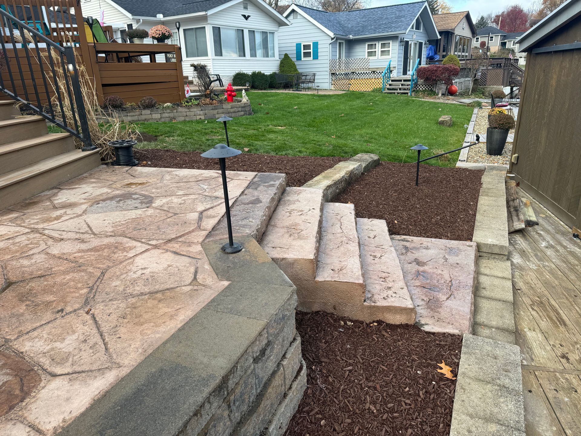 Stone patio steps beside a backyard lawn, with mulch beds and neighboring houses in view.
