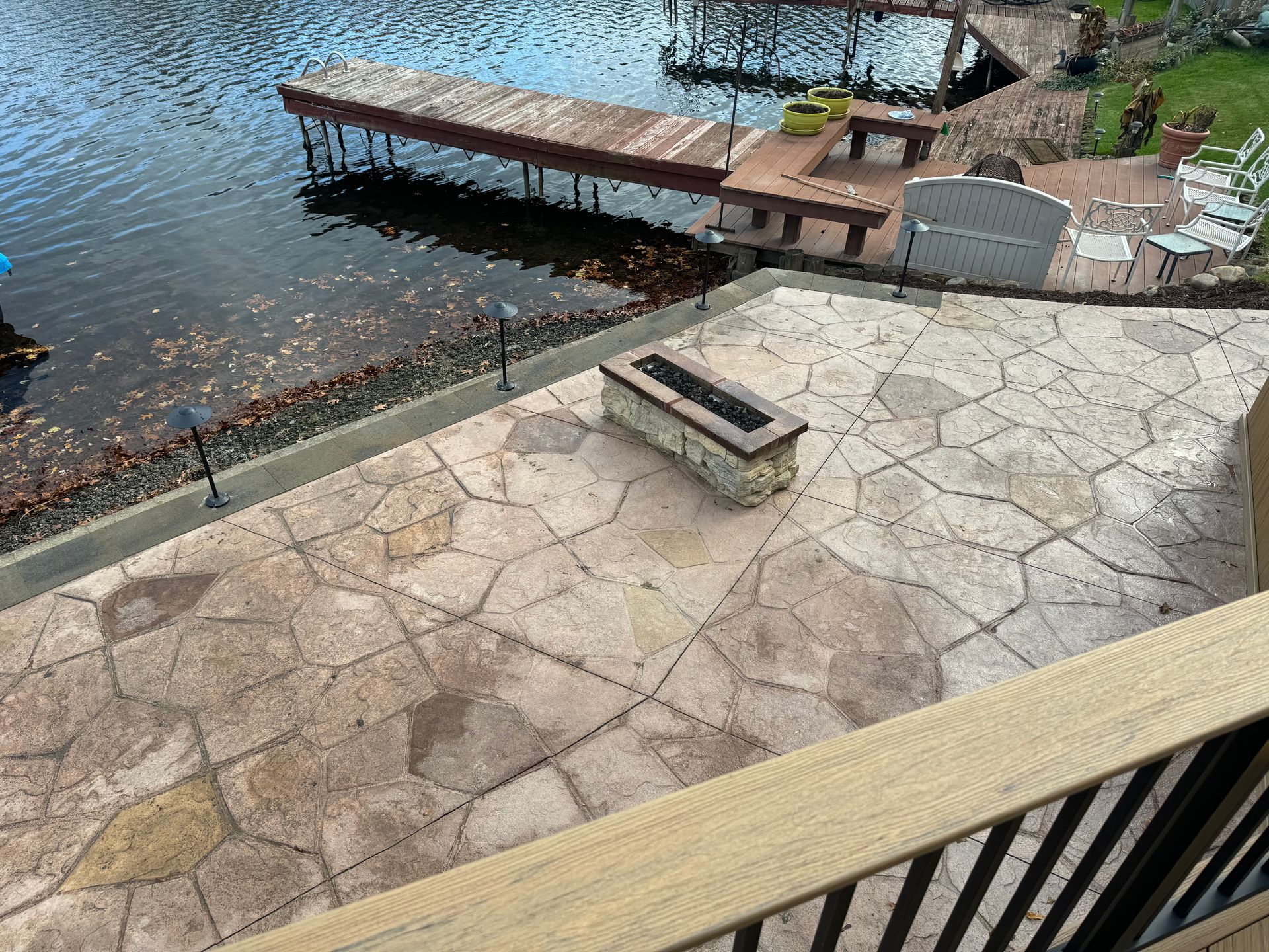 Stone patio beside a lake with a dock and fire pit, viewed from a balcony.