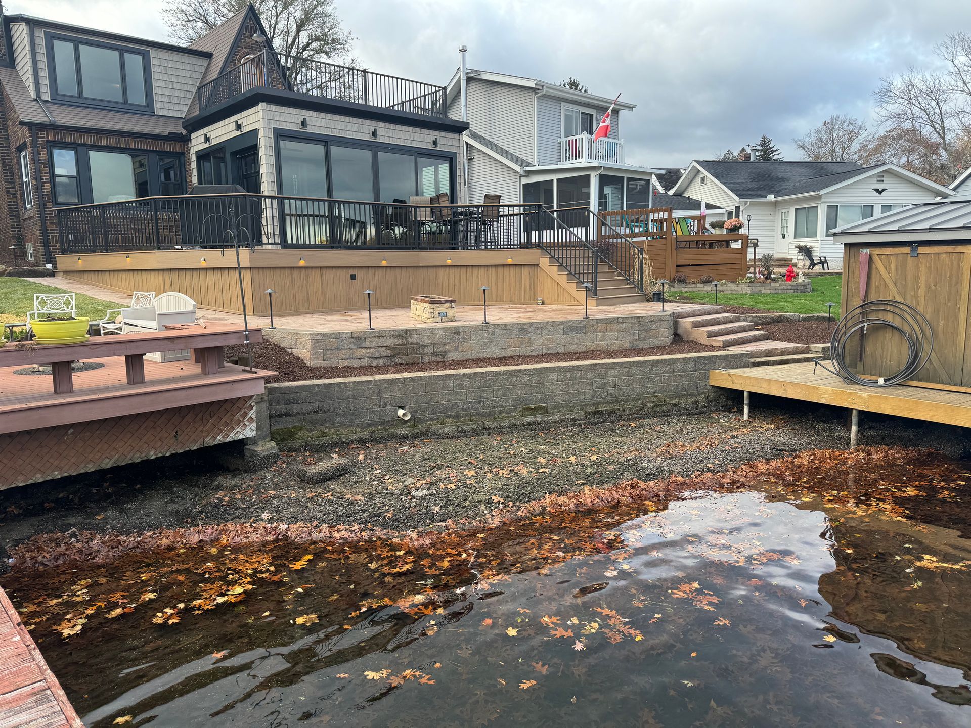 Backyard waterfront with deck, retaining wall, and houses under a cloudy sky
