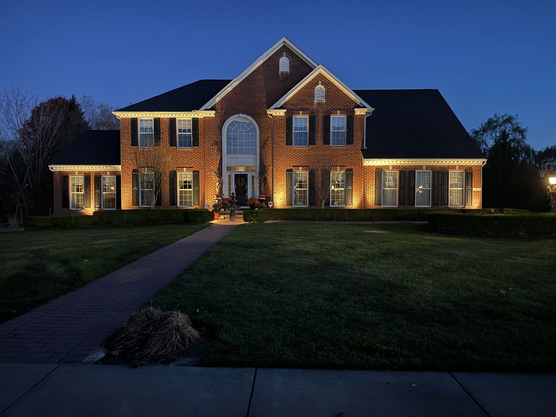 Large brick house with warm lights at dusk, front walkway, and a dark lawn.
