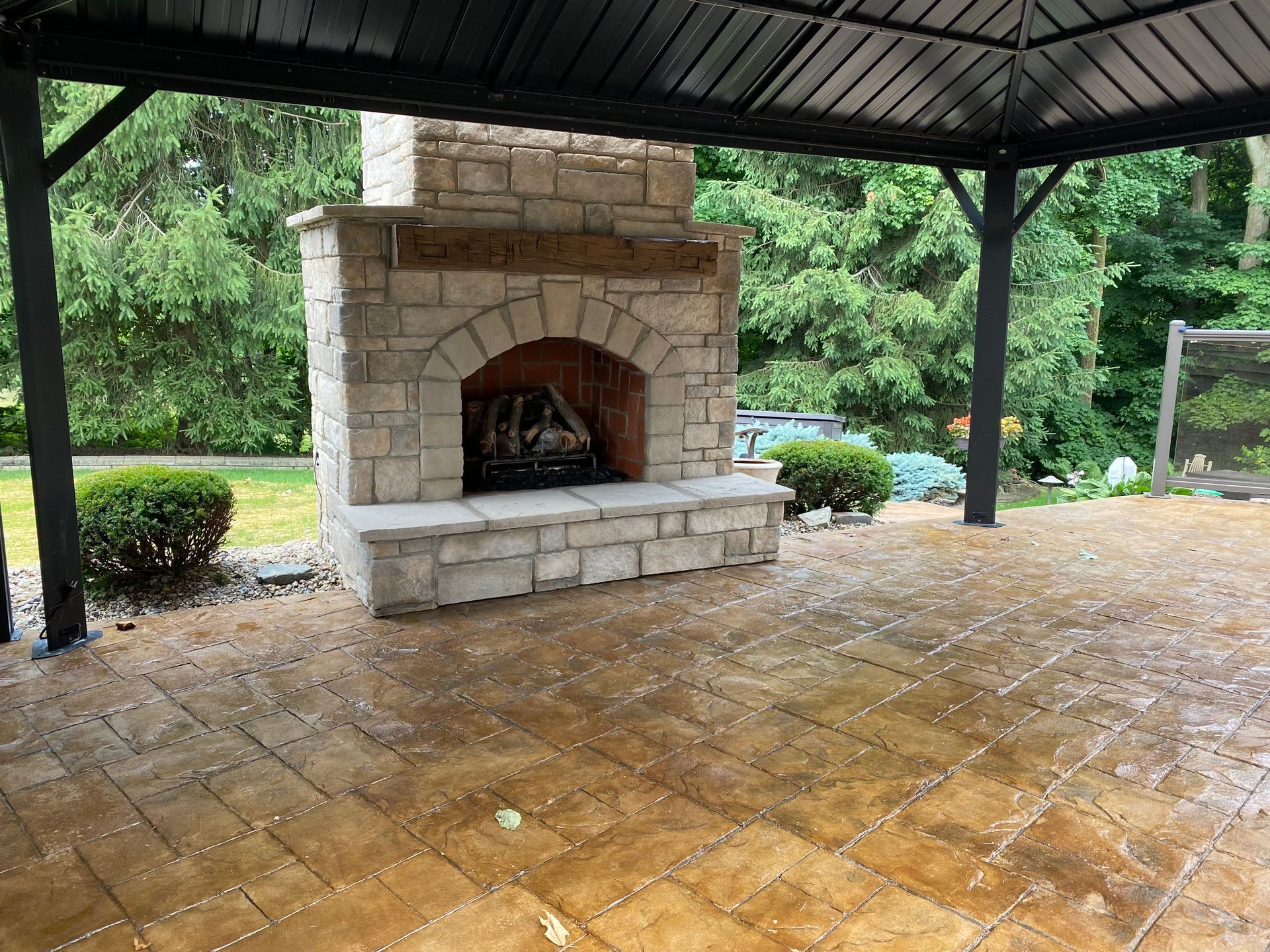 Covered outdoor stone fireplace on a wet patio under a metal roof, with green trees in the background.